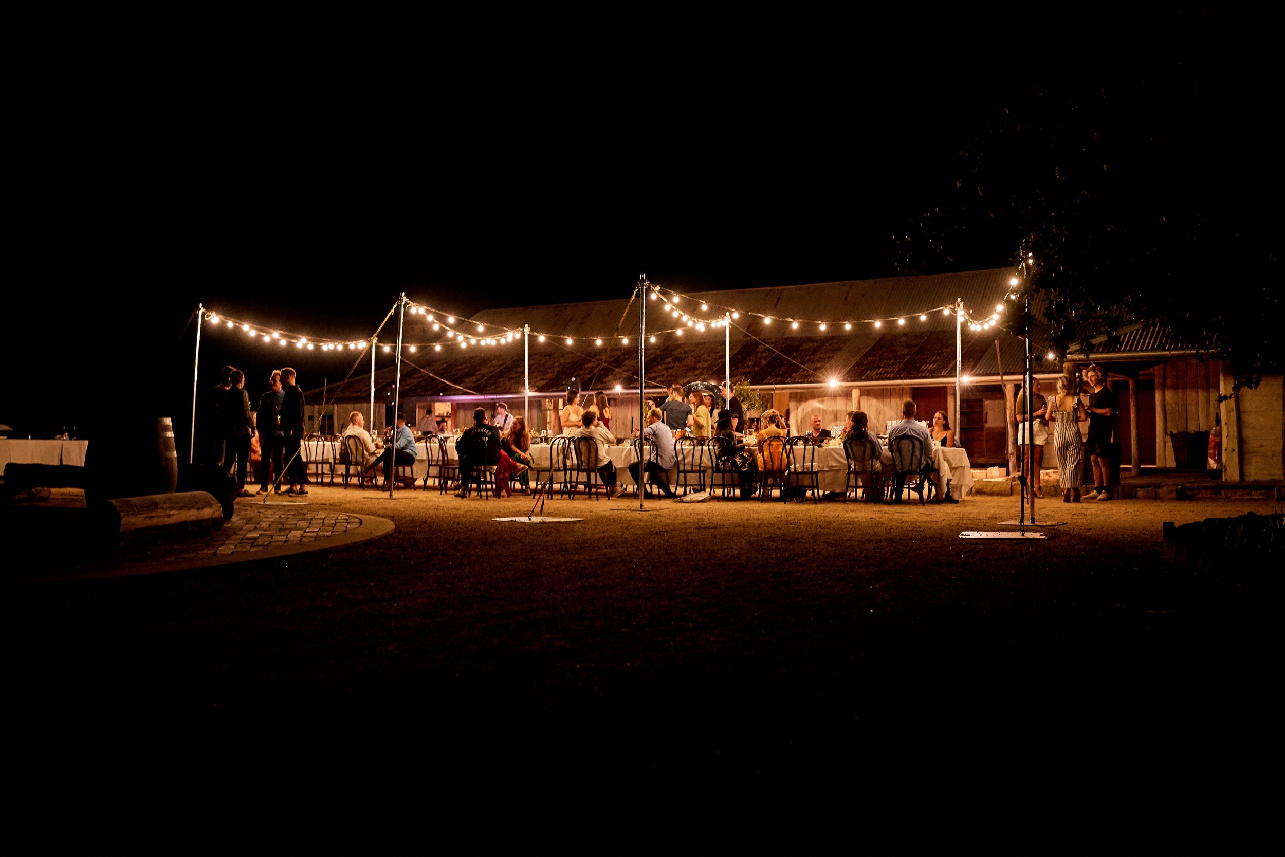 Outdoor evening gathering with string lights, tables, and people socializing beside a barn or similar building.