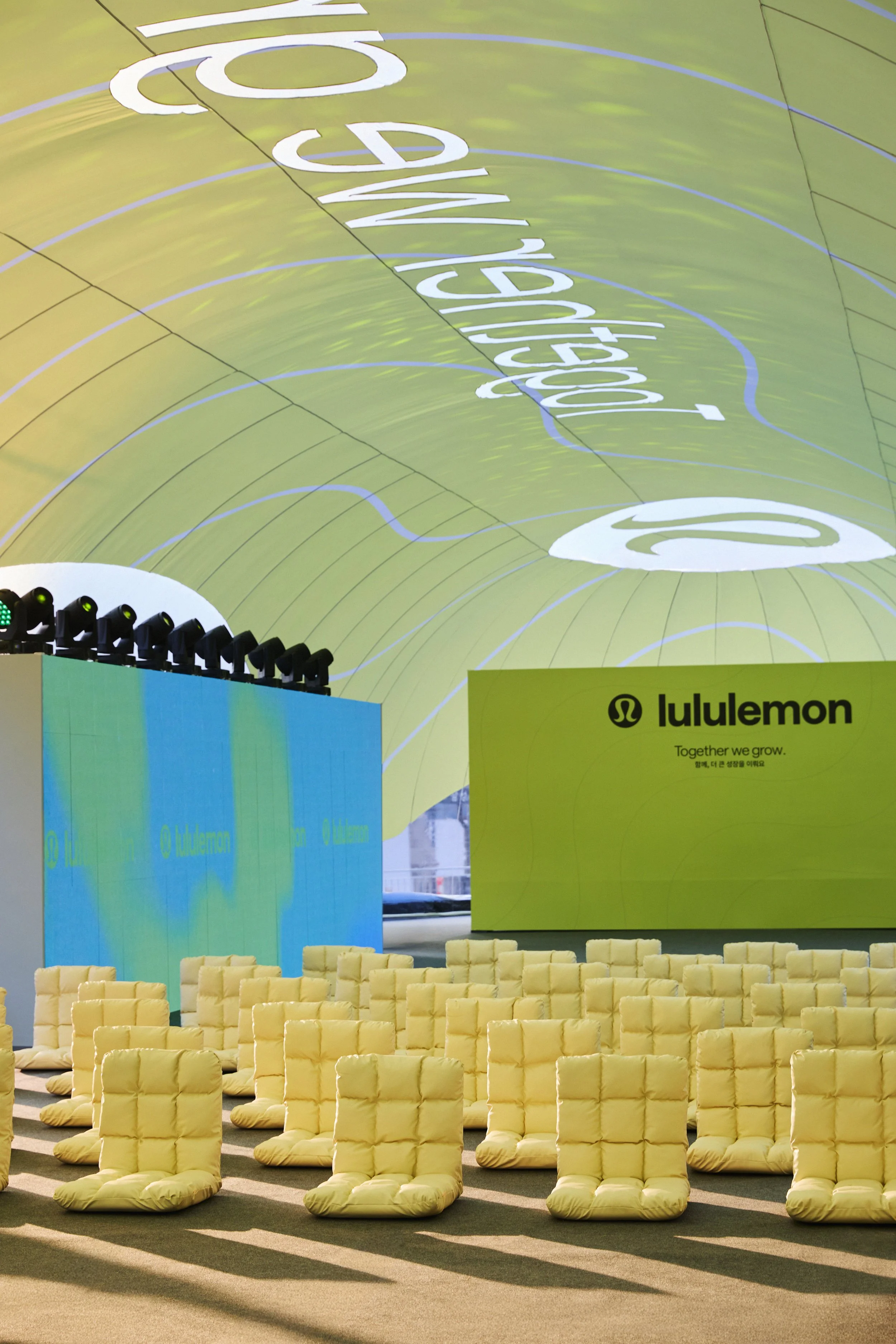 Empty yellow chairs arranged indoors with sunlight casting shadows on the floor. A large banner with the LuluLemon logo is visible in the background, along with a stage and lighting equipment.