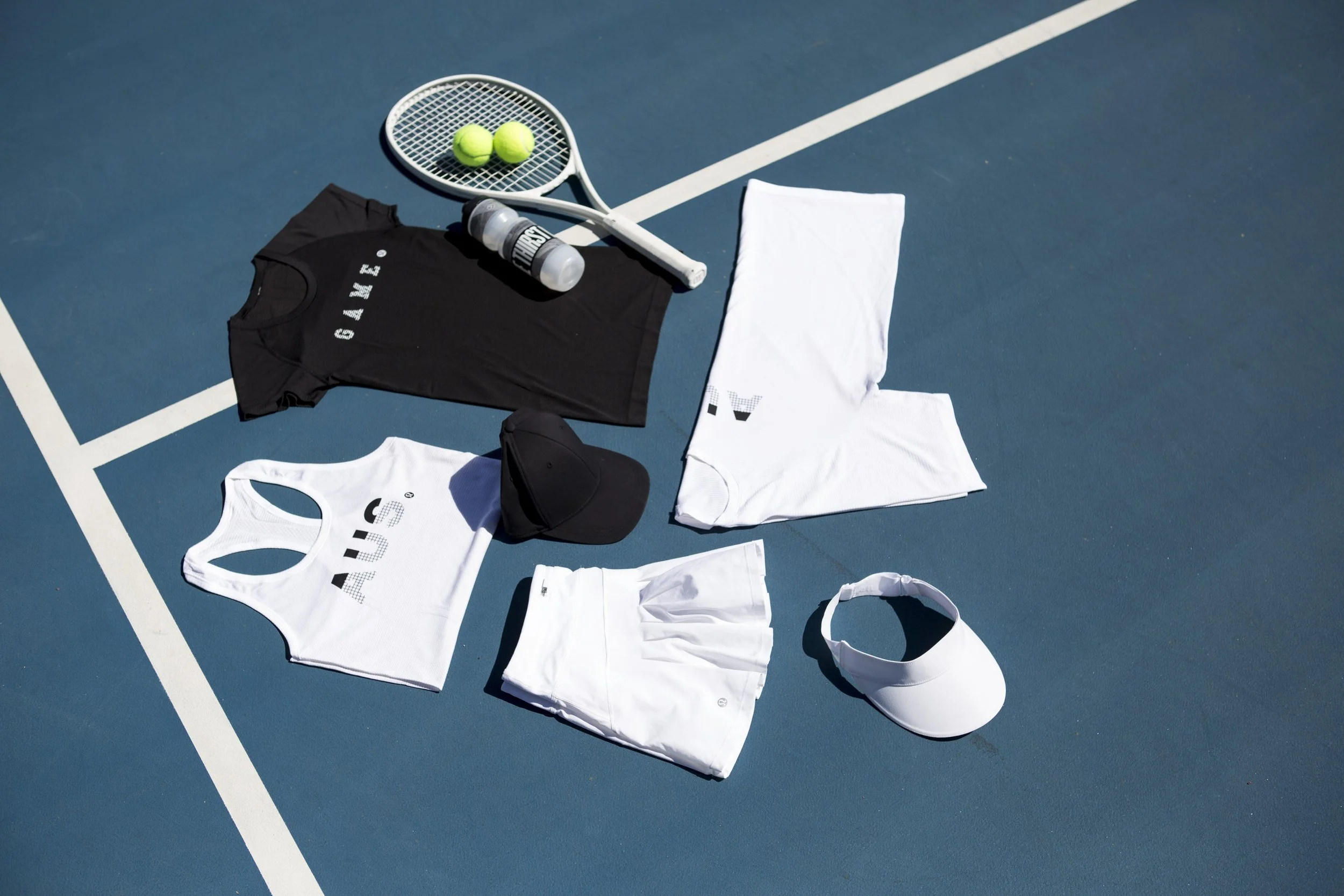 Tennis equipment and apparel arranged on a blue tennis court, including tennis balls, a racket, water bottle, cap, tank tops, and shorts.