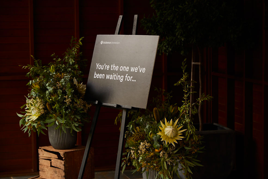 A sign with the Lululemon logo and the message "You're the one we've been waiting for..." surrounded by flower arrangements.