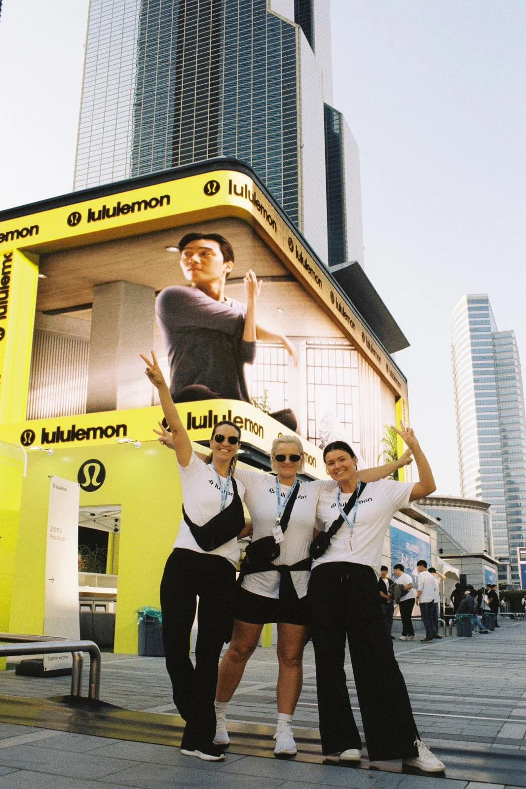 Three women wearing white T-shirts and black pants, standing in front of a large yellow booth with the 'lululemon' logo, smiling and posing with peace signs at an outdoor event in an urban area with tall buildings and a big digital billboard in the b