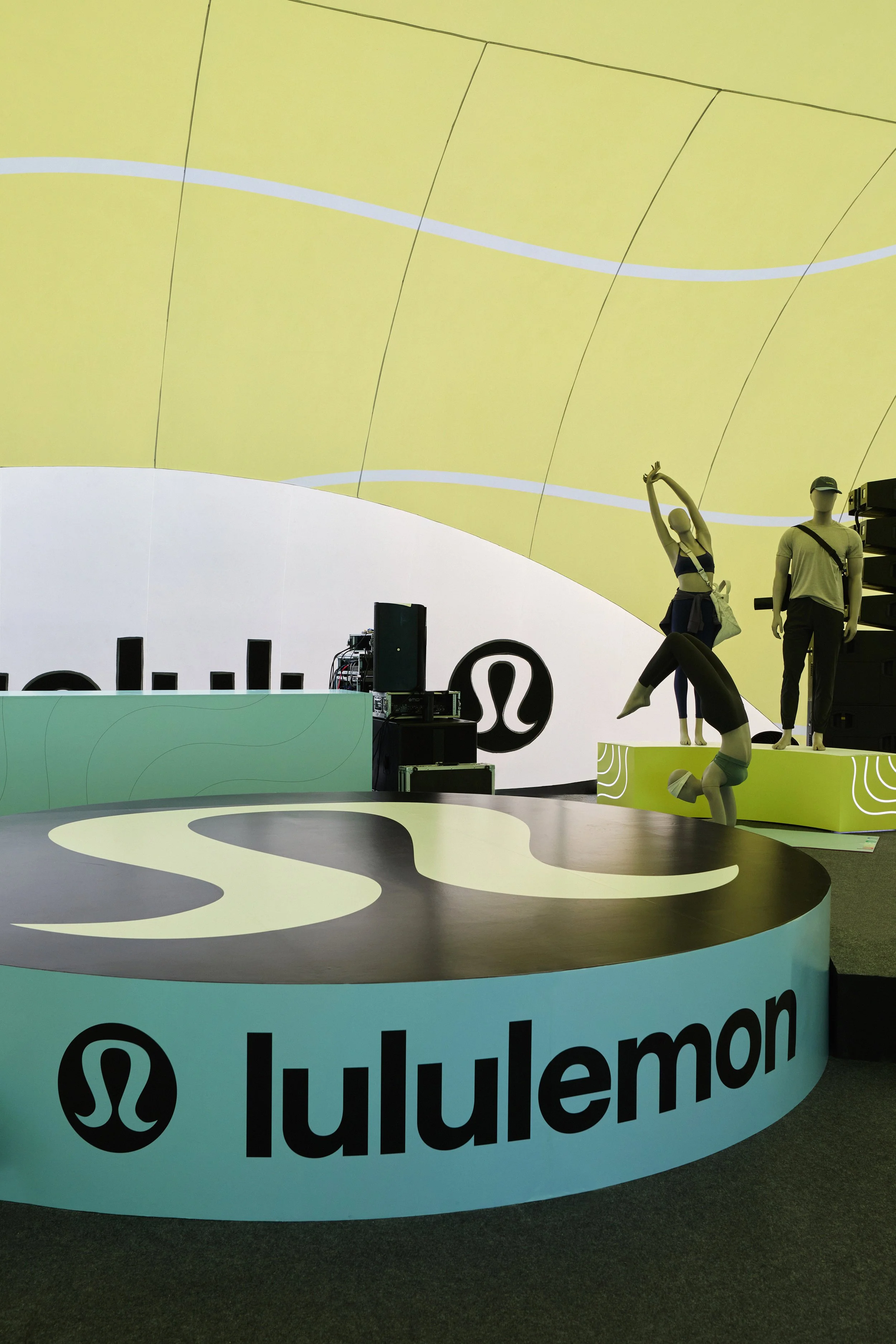 A large round display with the logo 'lululemon' and its symbol, featuring mannequins dressed in athletic clothing, with one mannequin doing a stretch pose and another standing, set against a yellow and white background.