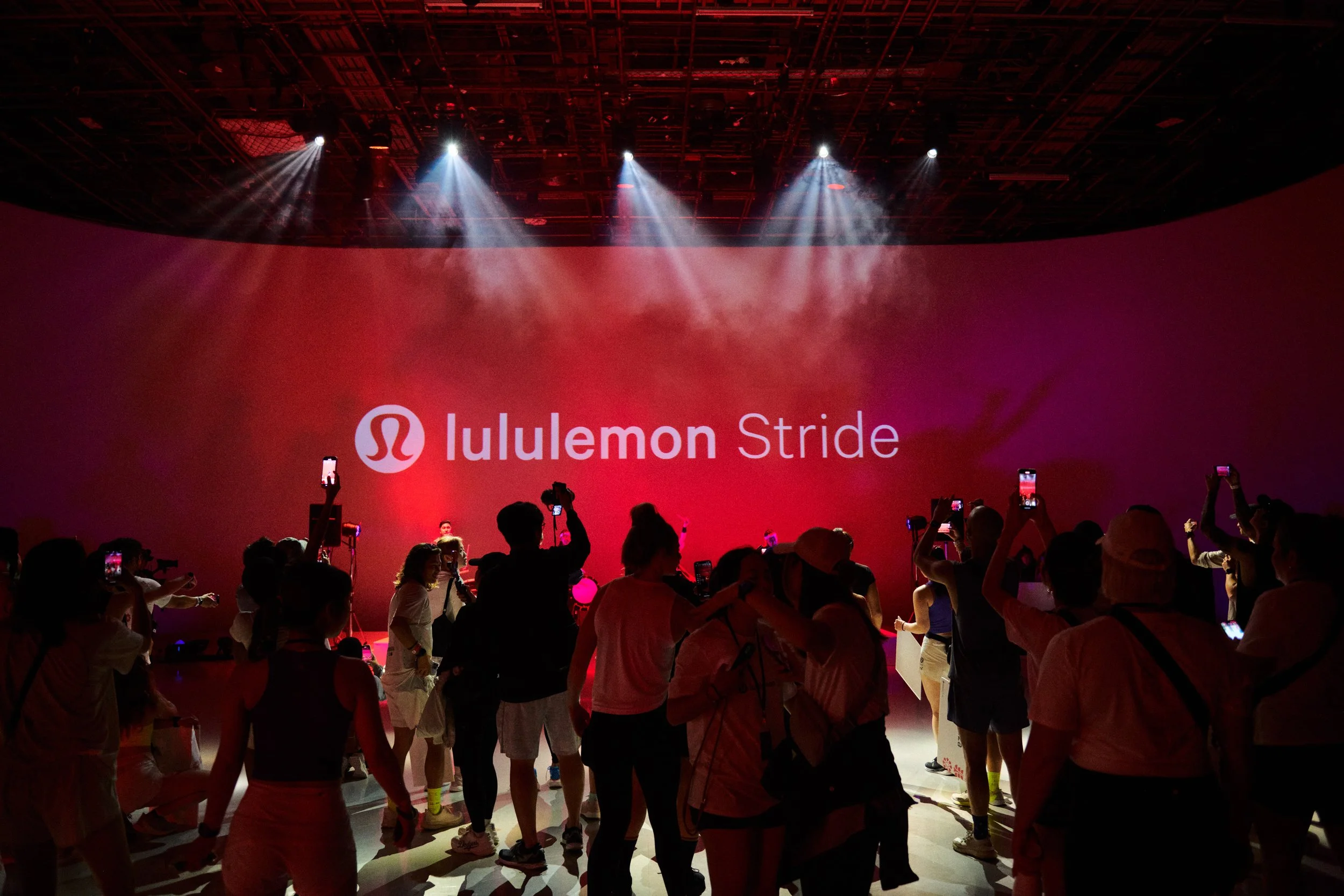 People gathered in a dark indoor space with a red background, standing and taking photos of a stage with spotlights and a large screen displaying the Luluemon Stride logo.