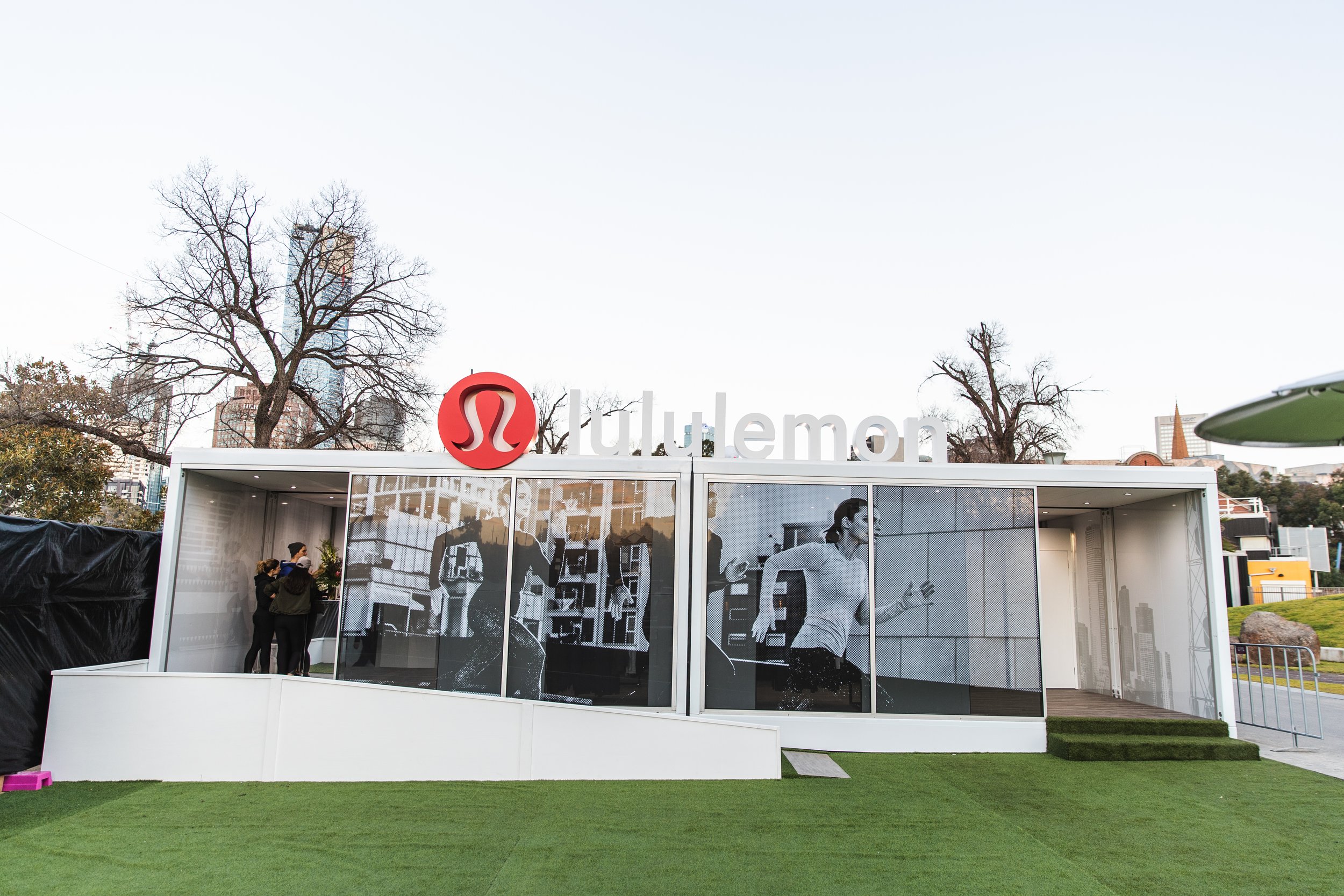A modern outdoor booth with the lululemon logo, featuring black and white images of people exercising, set on a grassy area with trees and city buildings in the background.