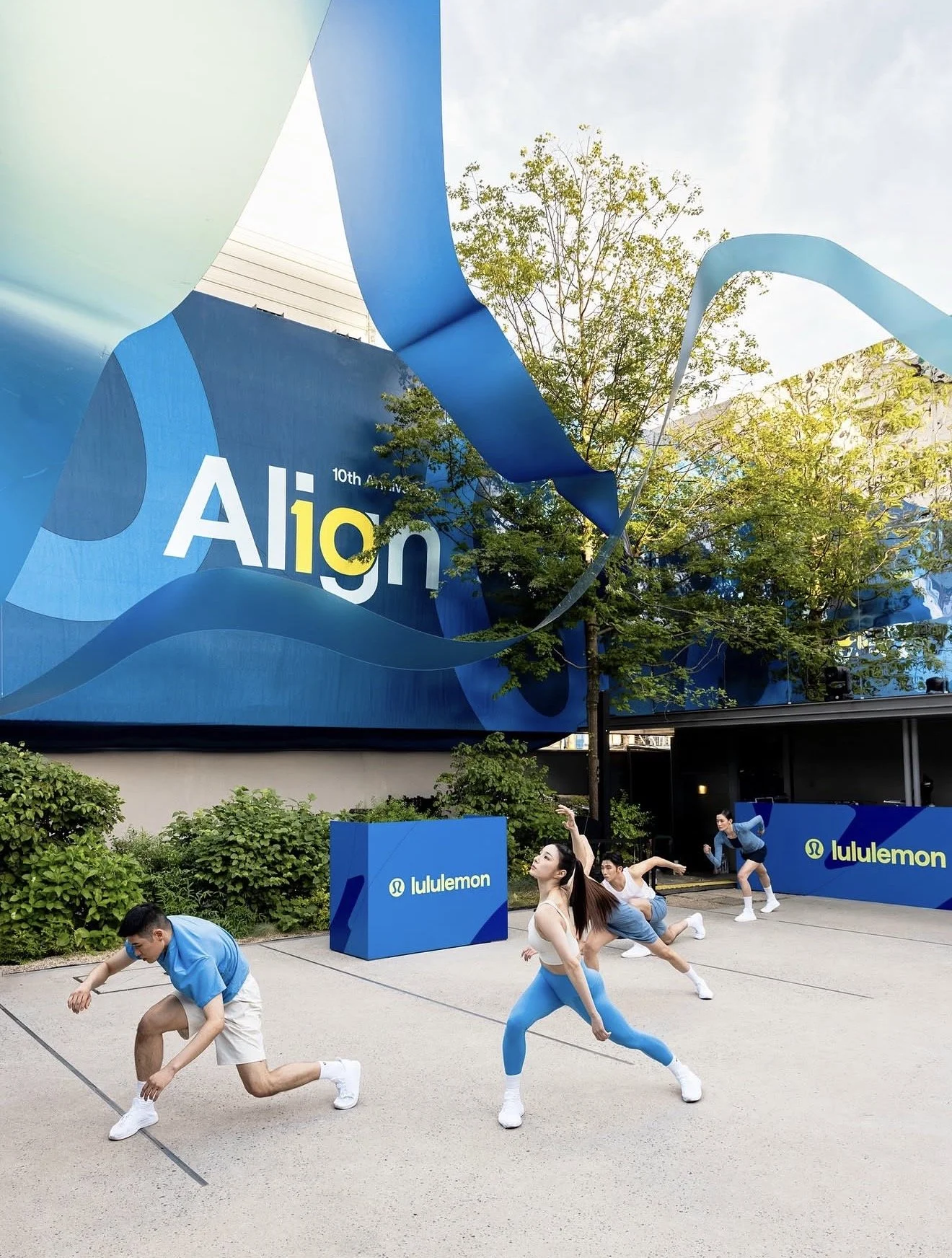Group of young people practicing yoga outdoors at a lululemon sponsored event, with a large blue and yellow event banner in the background.