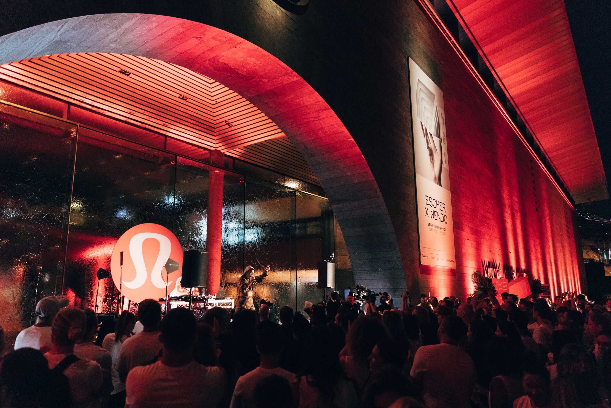 A large crowd of people gathered outdoors at night for a music event or concert. The stage is illuminated with red lighting and a DJ or performer is visible. The venue has an architectural structure with curved walls and a modern design.