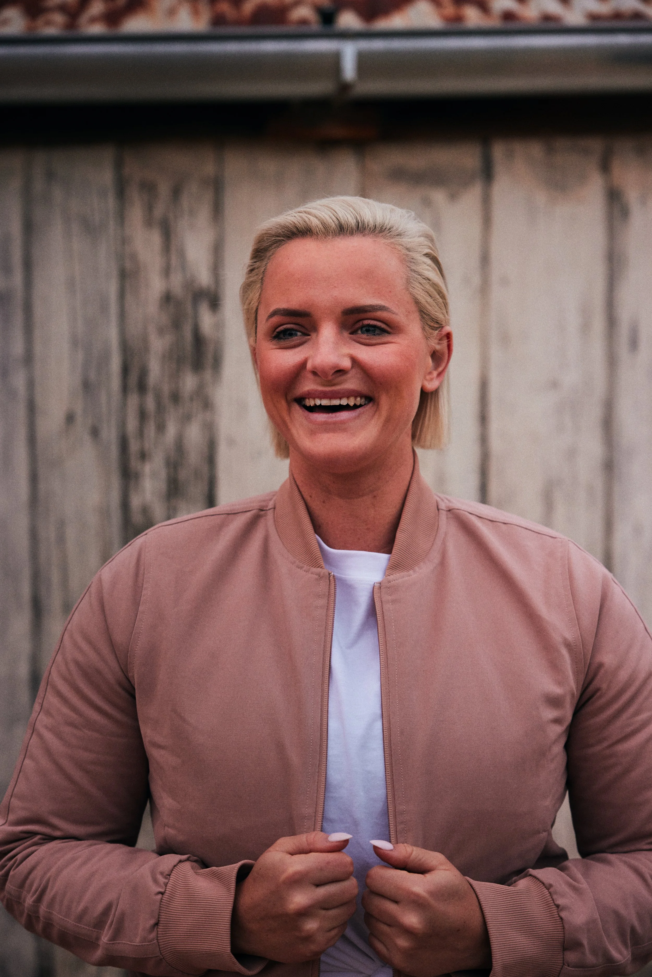 Woman with blonde hair smiling, wearing a light pink bomber jacket over a white shirt, standing in front of a rustic wooden background.