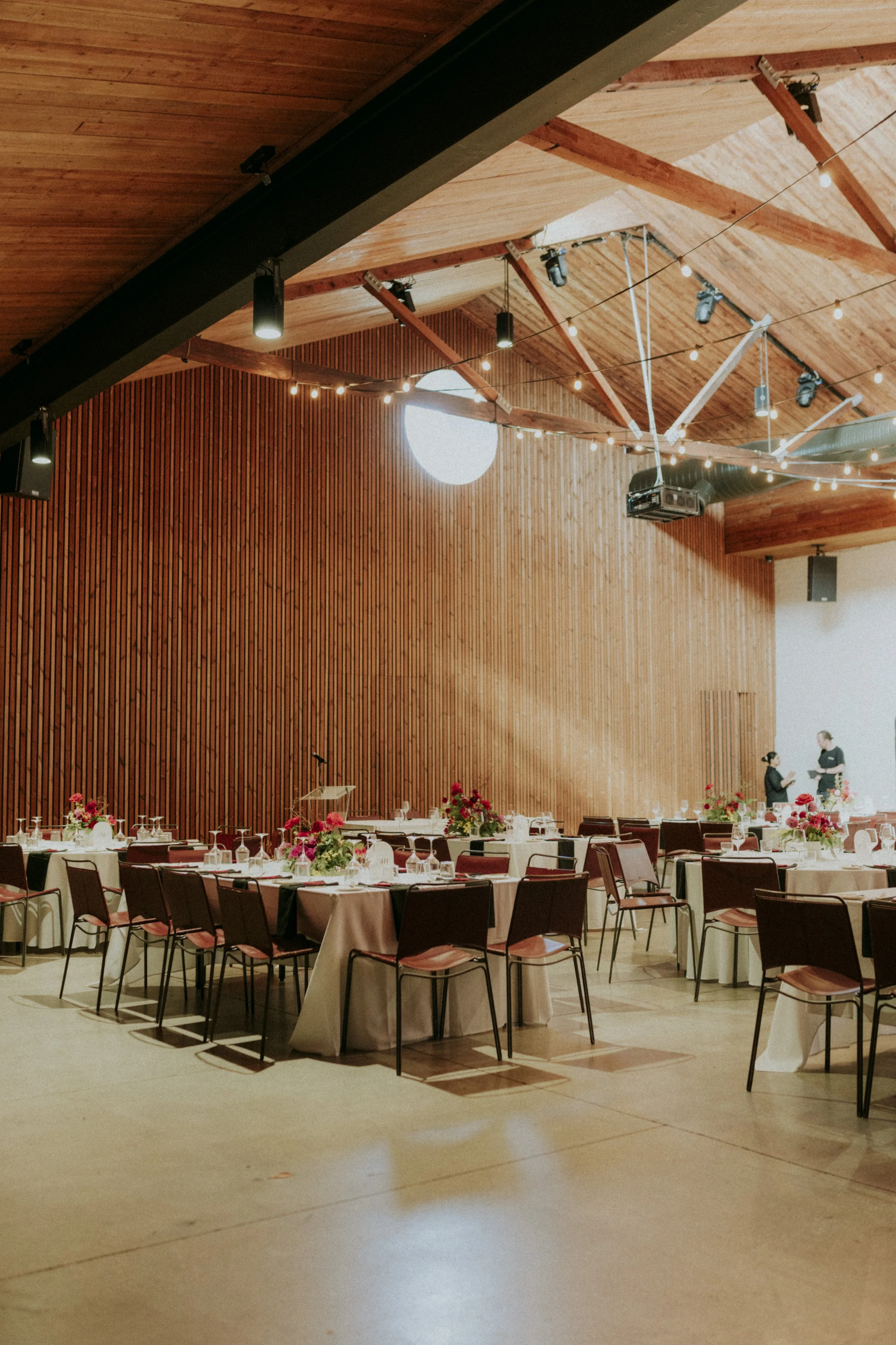 Elegant banquet hall with round tables covered in white tablecloths, decorated with pink and white floral centerpieces, and set with glassware and utensils. The room has a high wooden ceiling with exposed beams and string lights, and a large wooden p