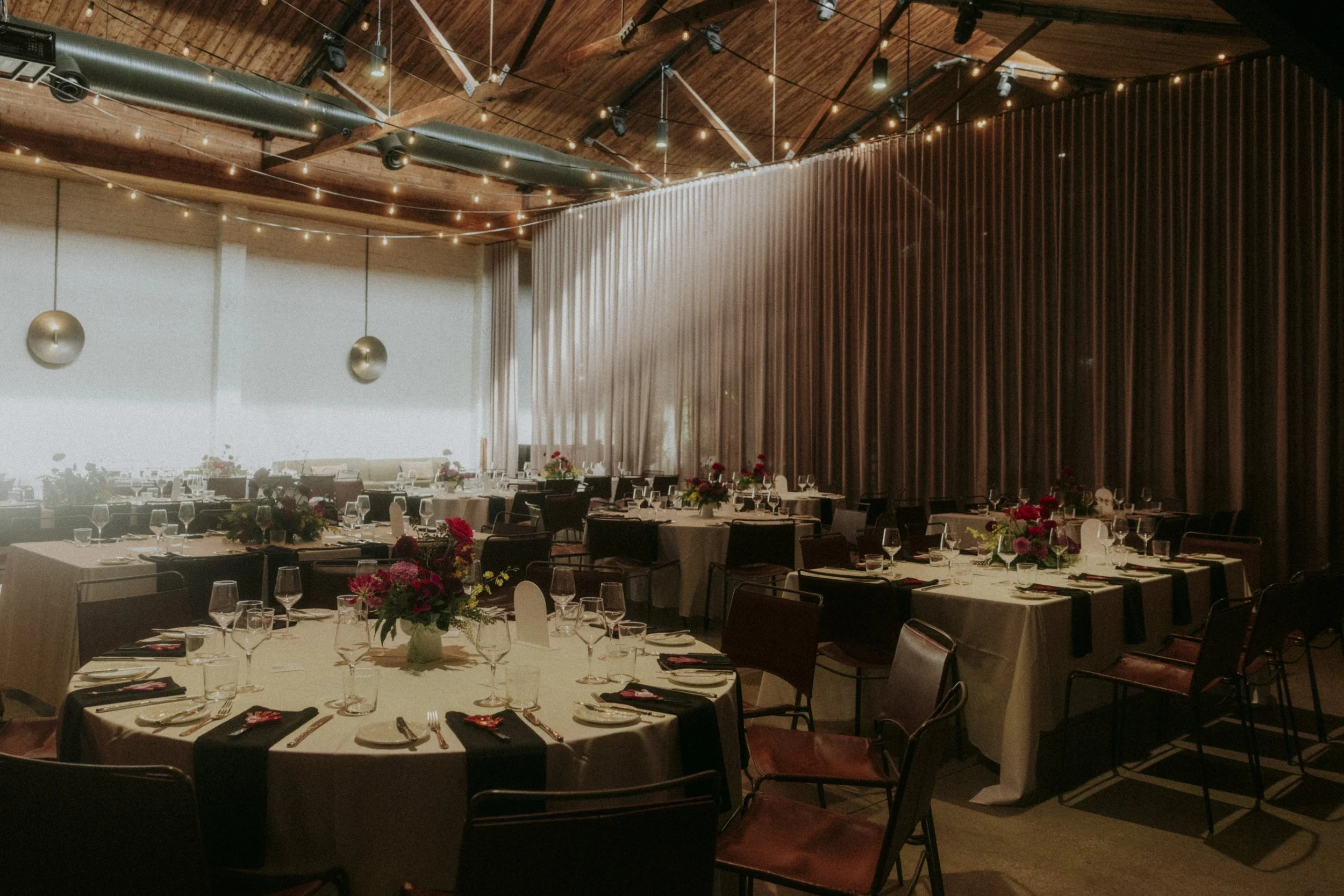 Elegant banquet room with round tables set with glassware, silverware, and napkins, decorated with vibrant floral centerpieces, wooden ceiling with string lights, and a large brown curtain dividing the space.