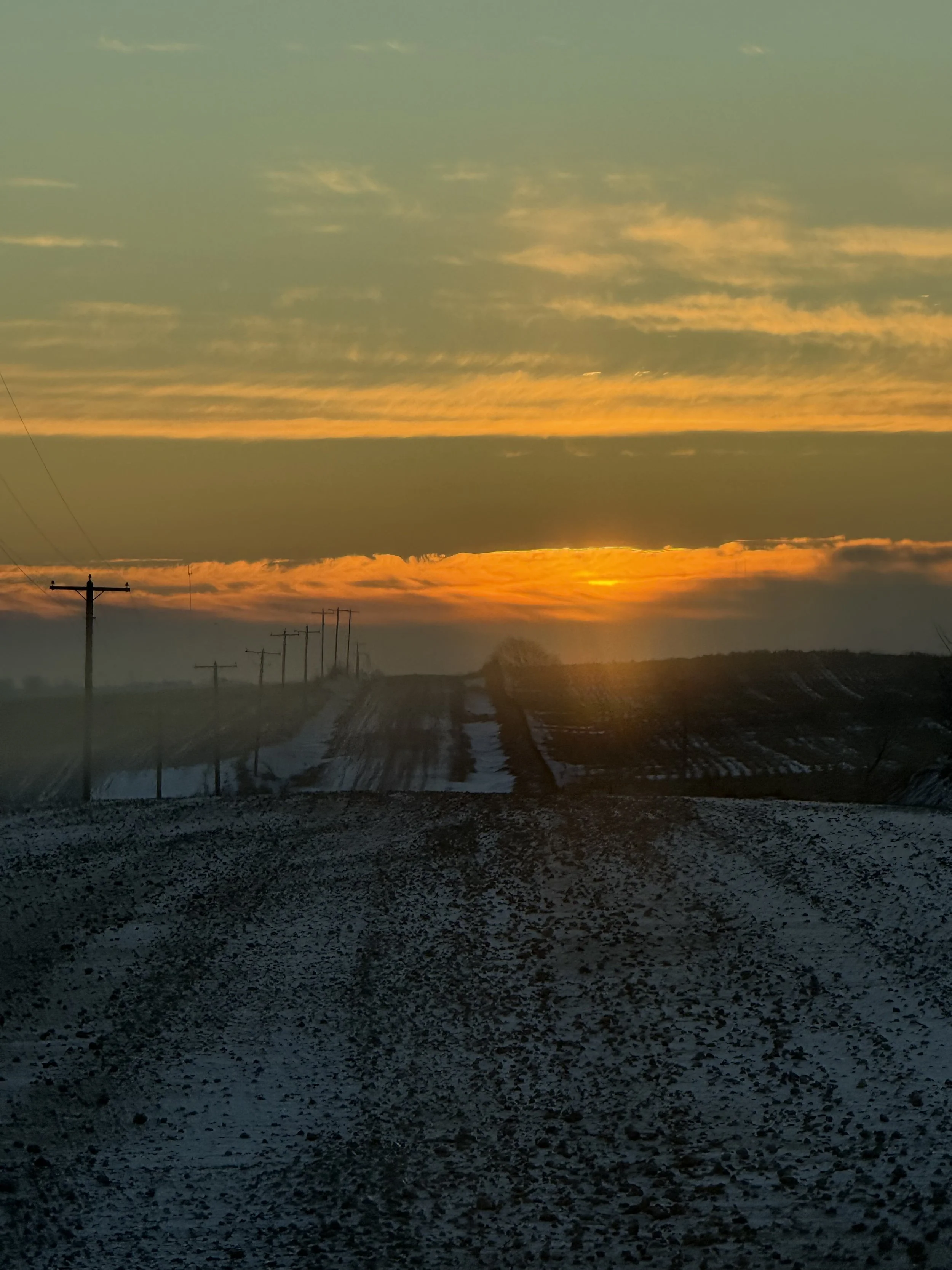 A rural dirt road during sunset, with utility poles on the left and fields on the right under a partly cloudy sky.