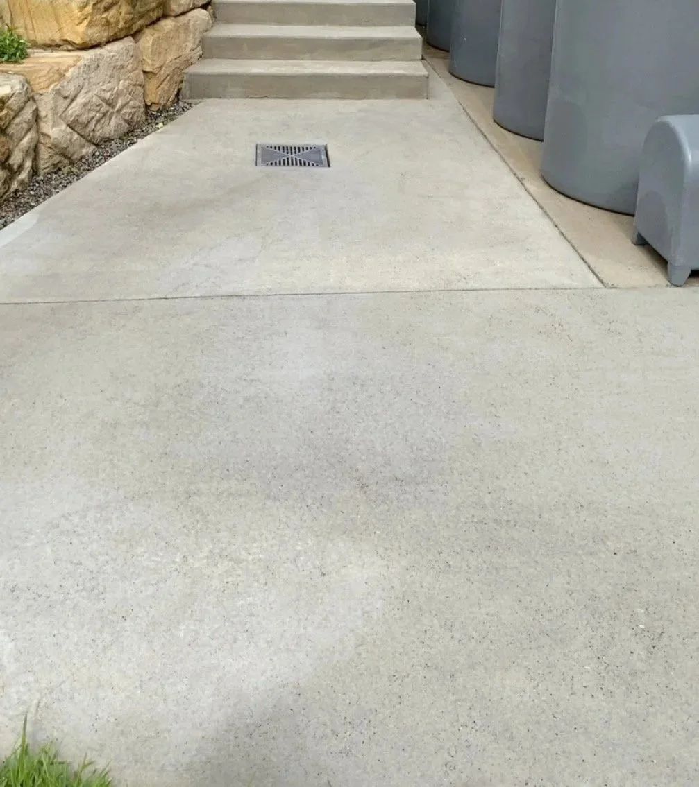 Concrete sidewalk leading to stairs with a drain in the middle, bordered by a stone wall on the left and trash cans on the right.