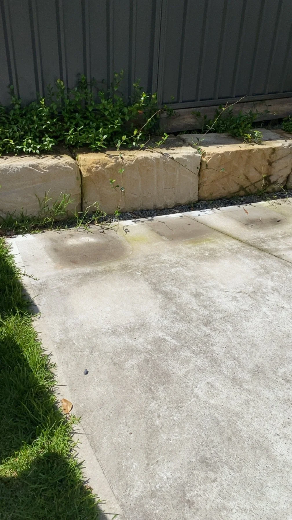 A concrete patio with visible stains and patches of moss, adjacent to a green lawn. Behind the patio, there is a raised flower bed bordered by large beige stone blocks, with green bushes and small plants growing in the bed. A gray metal fence is visible in the background.