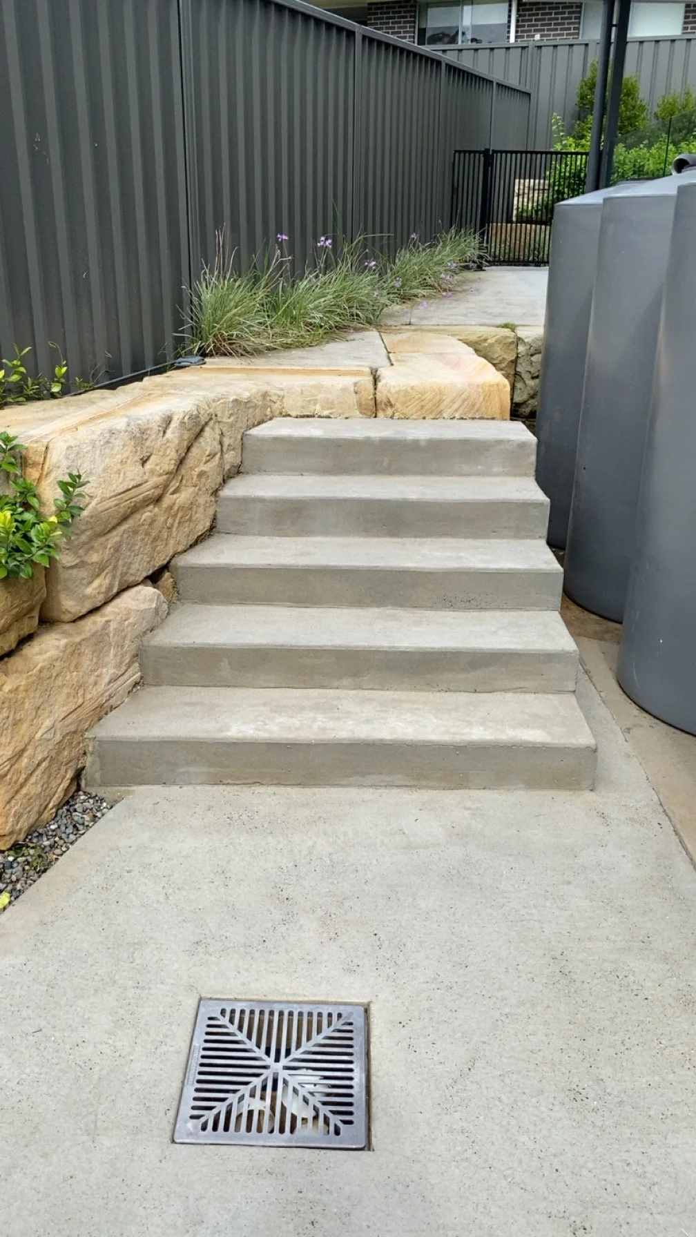 Concrete stairs leading up to a higher concrete pathway, bordered by a large rocky retaining wall with plants and purple flowers, with a gray metal fence and trash bins adjacent.