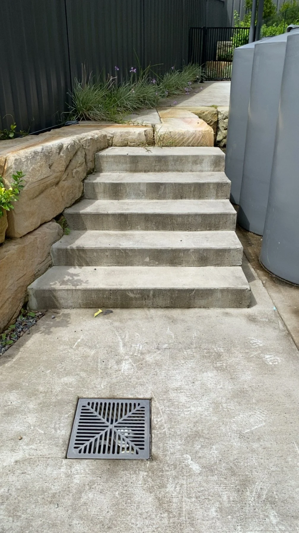 Concrete stairs leading up to a garden area with plants and rocks, located beside trash cans, with a drainage grate on the ground in the foreground.