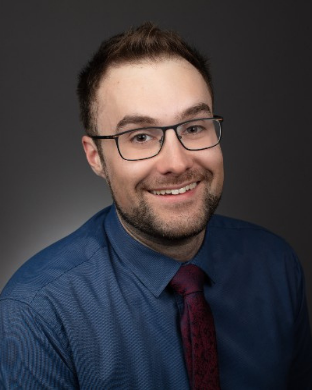 Portrait of Scott - Professional headshot of a smiling man wearing glasses, a blue dress shirt, and a maroon tie against a dark background.