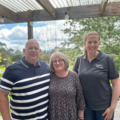 Three people standing outdoors under a wooden pergola, smiling at the camera. One man on the left wearing a navy polo with white stripes, a woman in the middle with shoulder-length hair and glasses, wearing a floral blouse, and a young woman on the right with a ponytail, wearing a dark gray polo shirt with a logo. There are trees and a cloudy sky in the background.