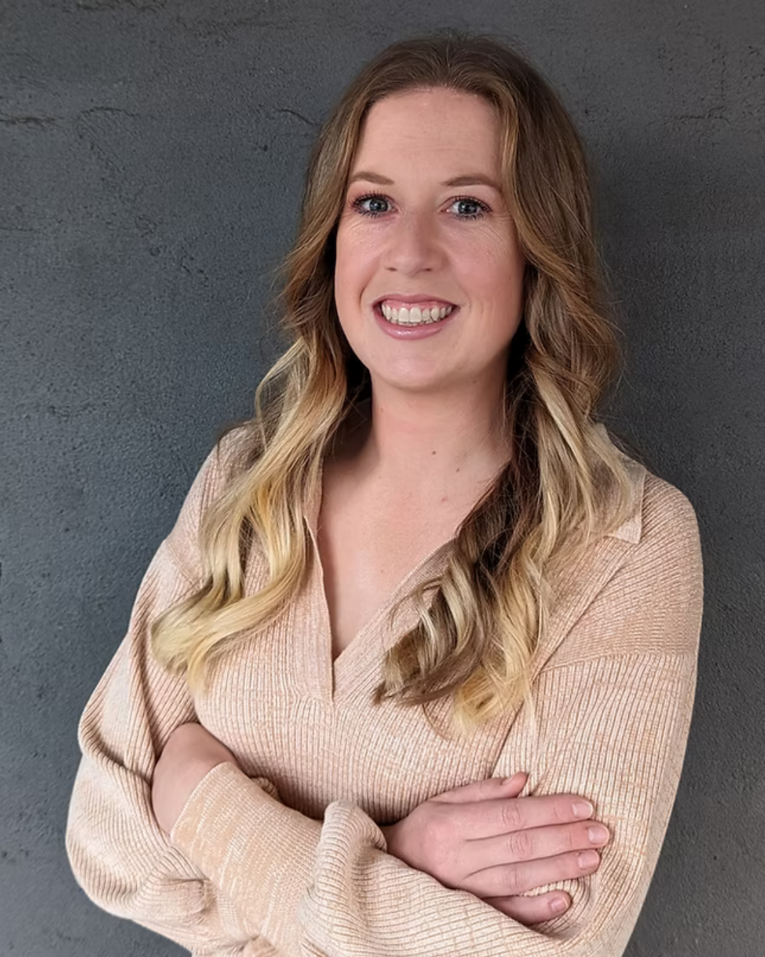 Portrait of Elleesha - A woman with long, blonde, wavy hair, smiling with arms crossed, wearing a beige ribbed sweater, standing against a dark gray textured wall.