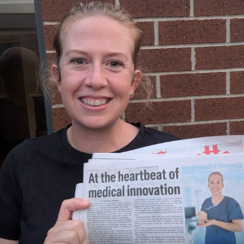 A young woman with fair skin, red hair, smiling, holding a stack of newspapers with the headline 'At the heartbeat of medical innovation,' standing in front of a brick wall.