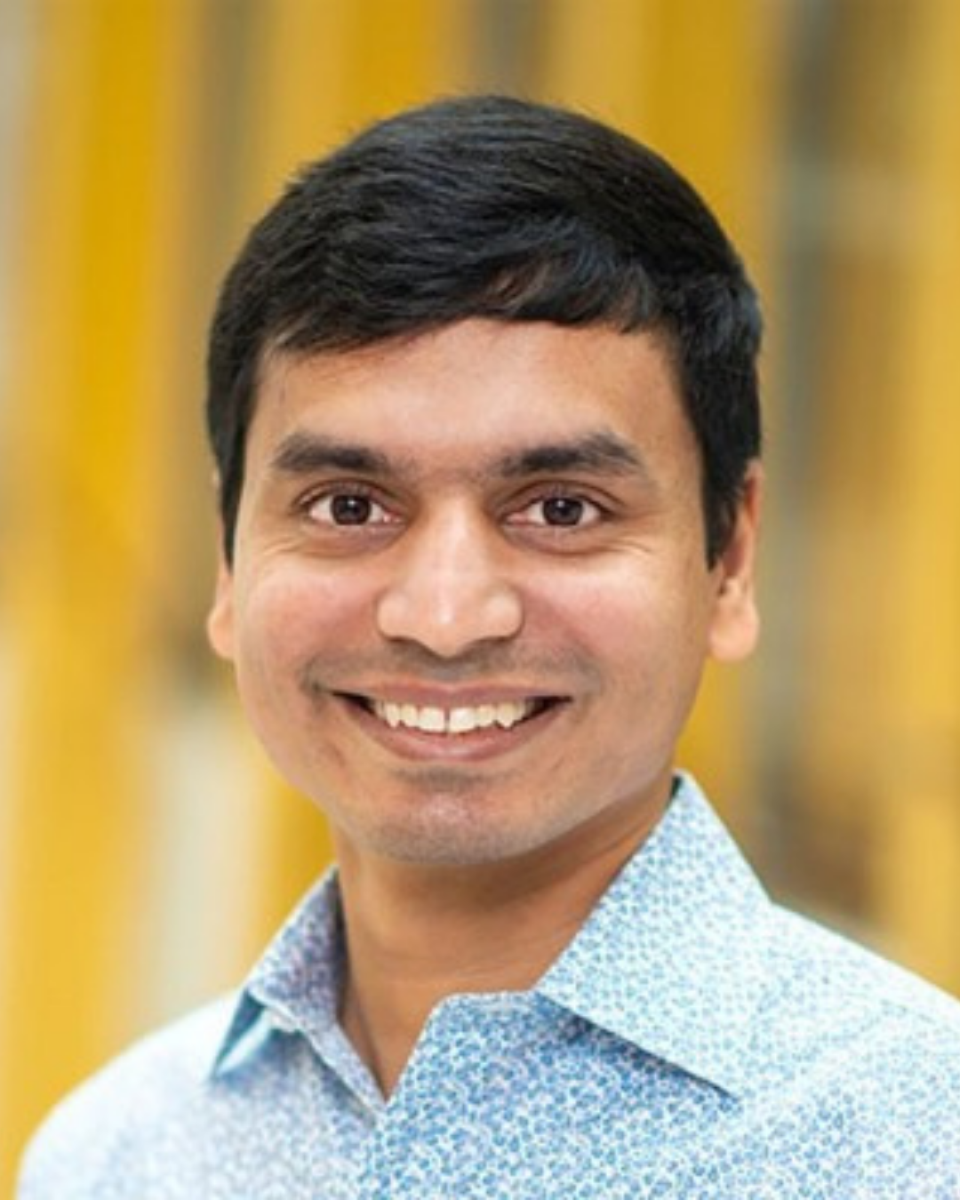 Portrait of Prof. Sharath Sriram - A smiling man with short black hair wearing a light blue patterned shirt, indoors with a yellow background.