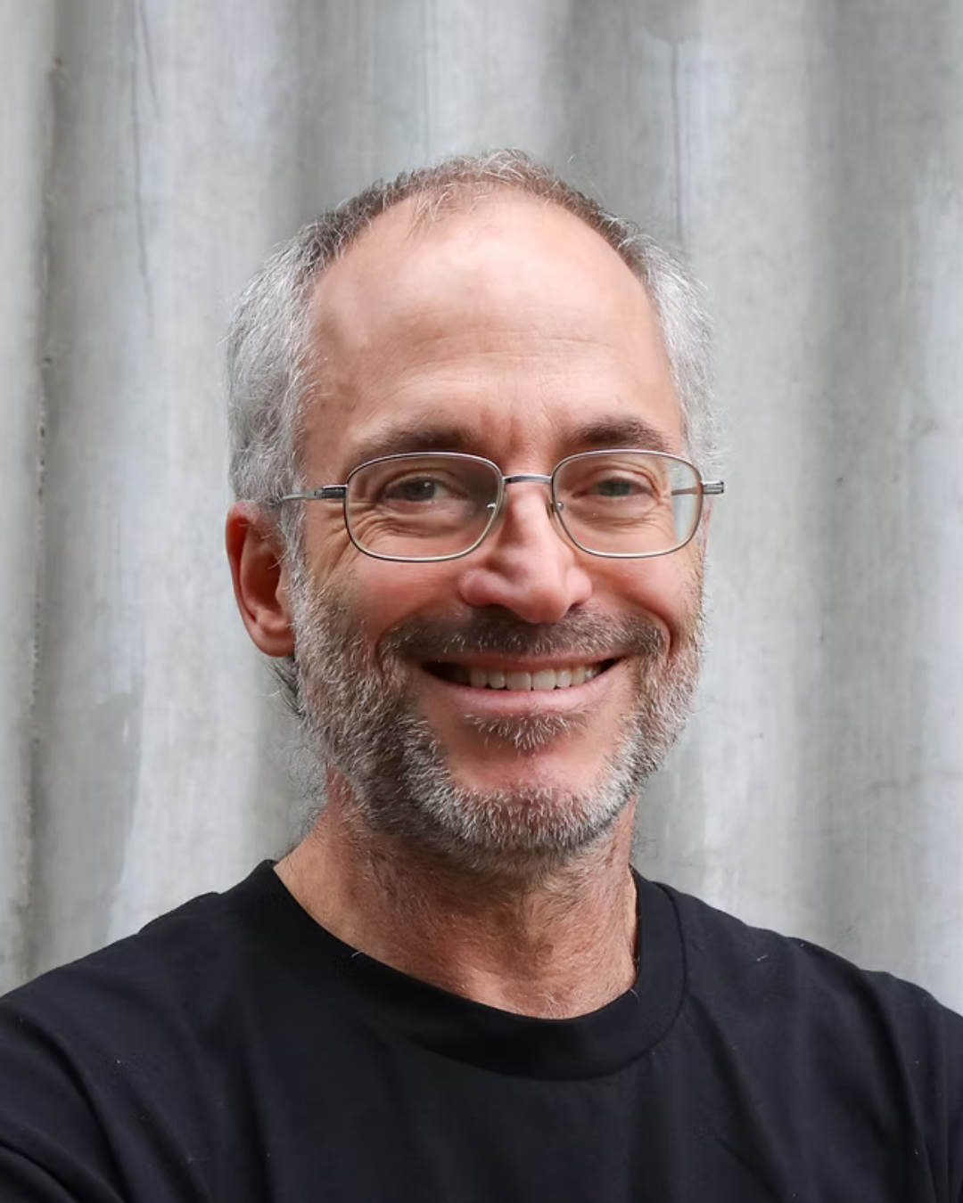 Portrait of Paul - A smiling middle-aged man with gray hair, beard, and glasses, wearing a black t-shirt, standing in front of a light-colored curtain.