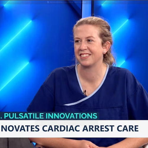 A woman in medical scrubs being interviewed on a television set with a blue background. There is text on the screen about cardiac arrest care innovation.