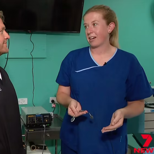 A female medical professional in blue scrubs speaking to a man in a hospital room, with medical equipment and a television on the wall in the background.