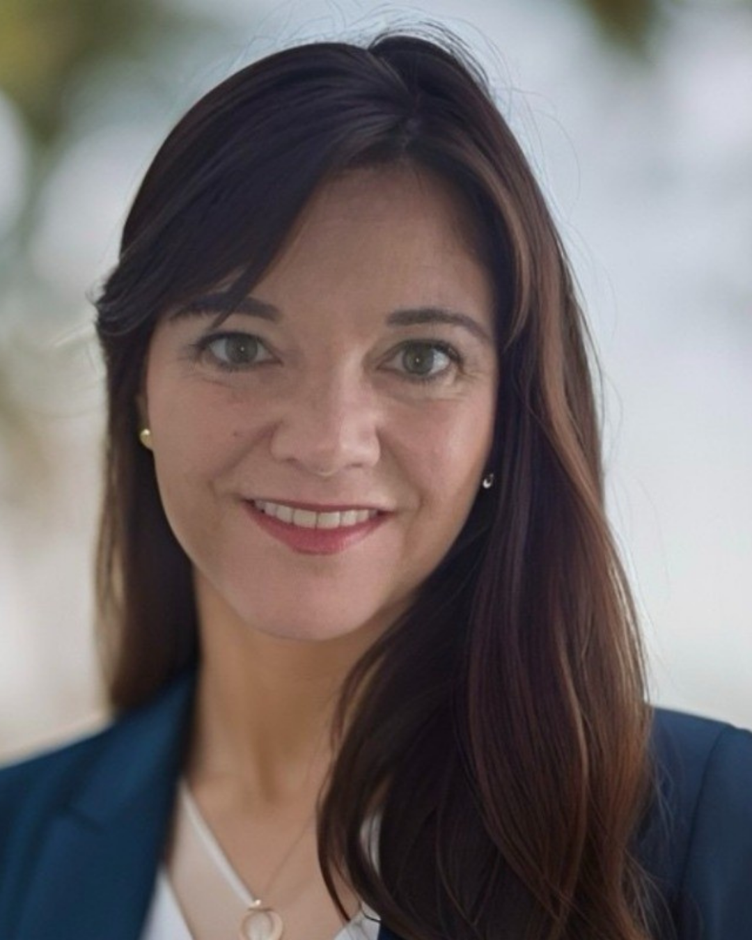 Portrait of Nicolette - A woman with long brown hair, gray eyes, and a friendly smile, wearing a blue blazer and pearl earrings, standing outdoors.