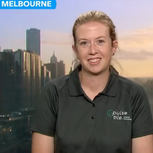 A woman with blonde hair pulled back, wearing a black polo shirt with a logo, is smiling during a news interview with a city skyline and a sunset sky in the background.