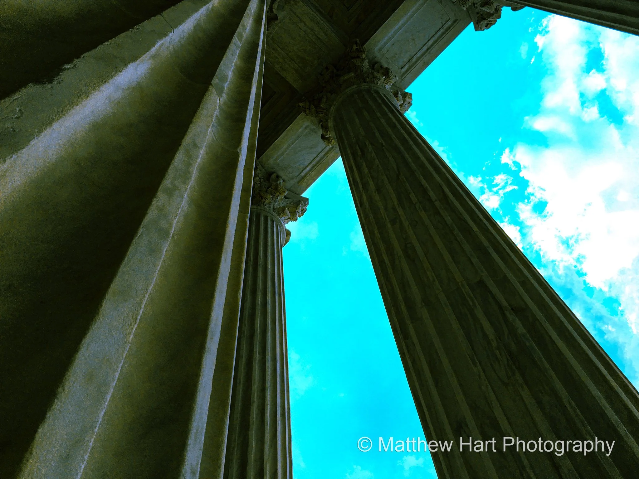 Fine art photograph looking up between tall fluted stone columns toward a bright blue sky with clouds in Washington, D.C.