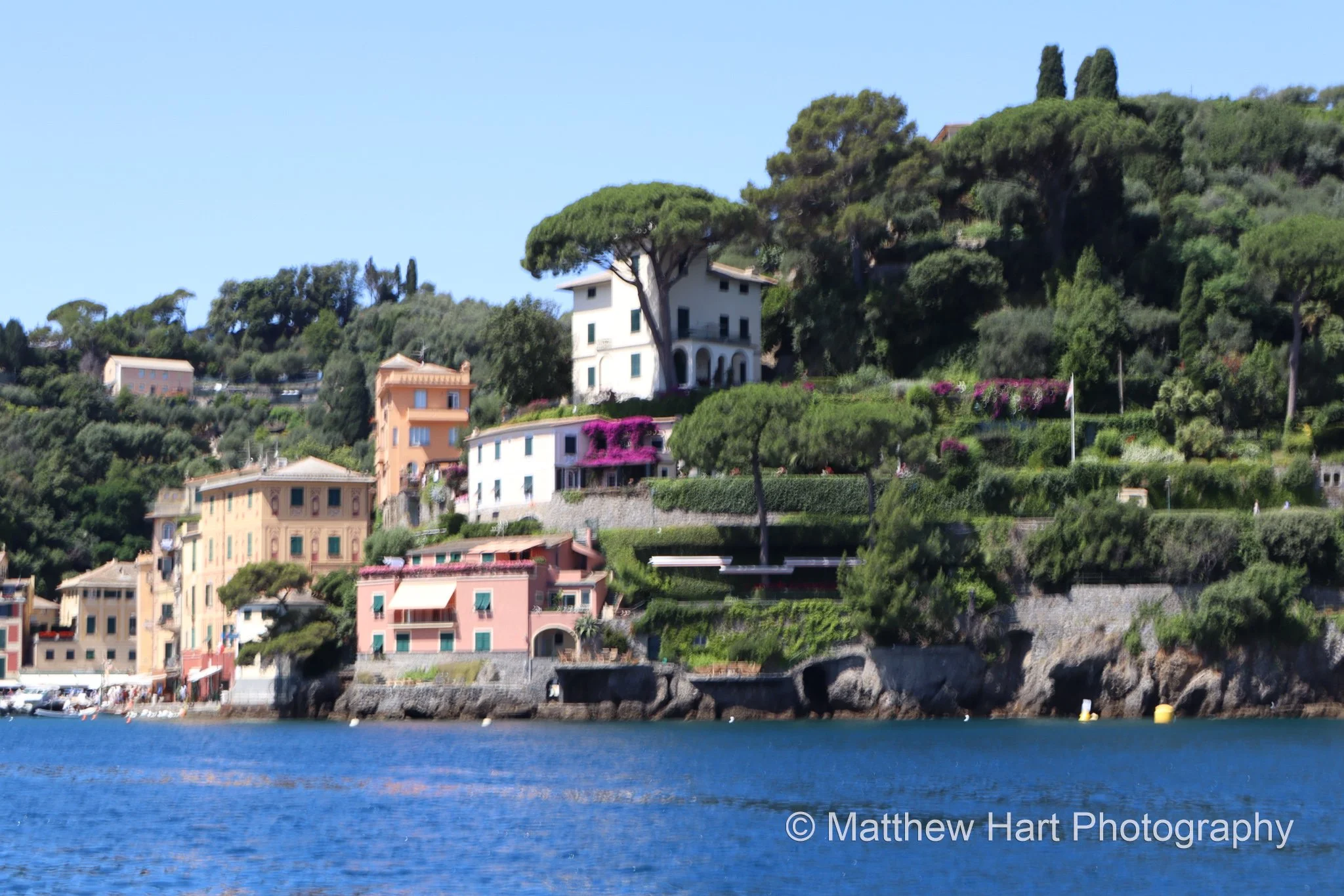 Fine art photograph of colorful houses at the water's edge in a green, pine-covered cove on the Italian Riviera.