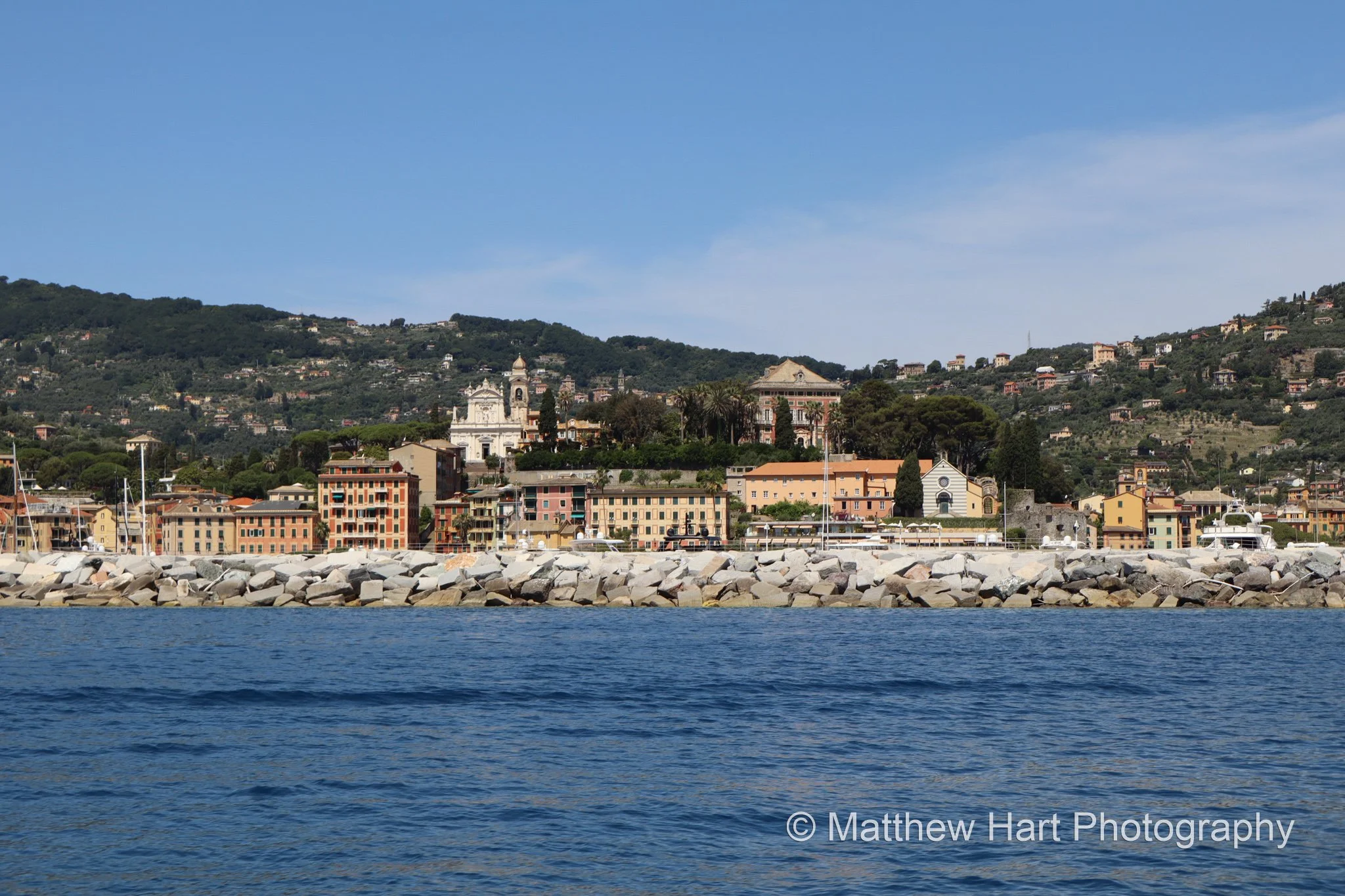 Fine art photograph of Santa Margherita Ligure, Italy, seen from the sea behind a stone breakwater, with a white church façade and green hills.