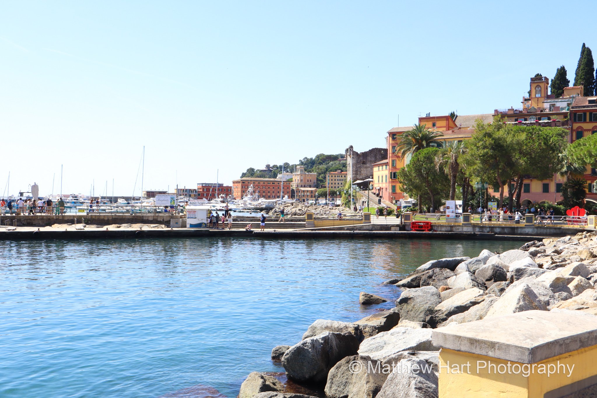Fine art photograph of the harbor at Santa Margherita Ligure, Italy, with breakwater boulders, calm water, and colorful buildings beyond the marina.