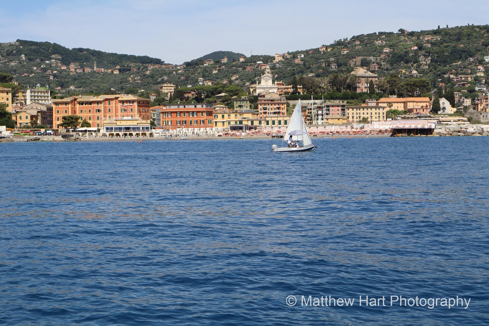Fine art photograph of a white sailboat on a blue bay in front of a pastel seafront town and wooded hills on the Italian Riviera.