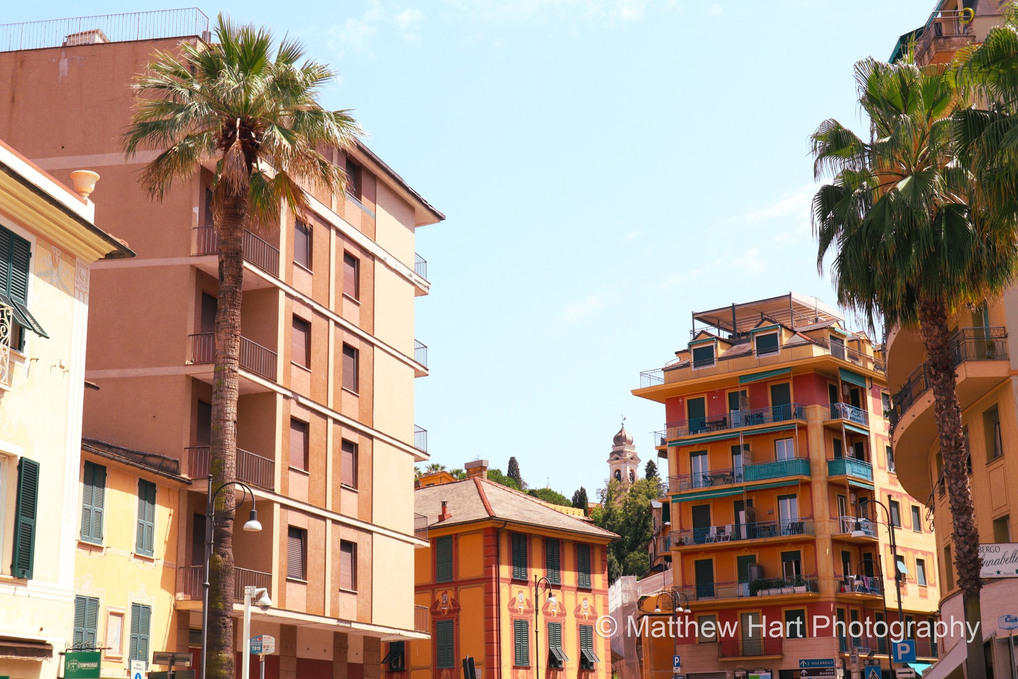 Fine art photograph of tall palm trees between apricot and coral apartment buildings with balconies in Santa Margherita Ligure, Italy.