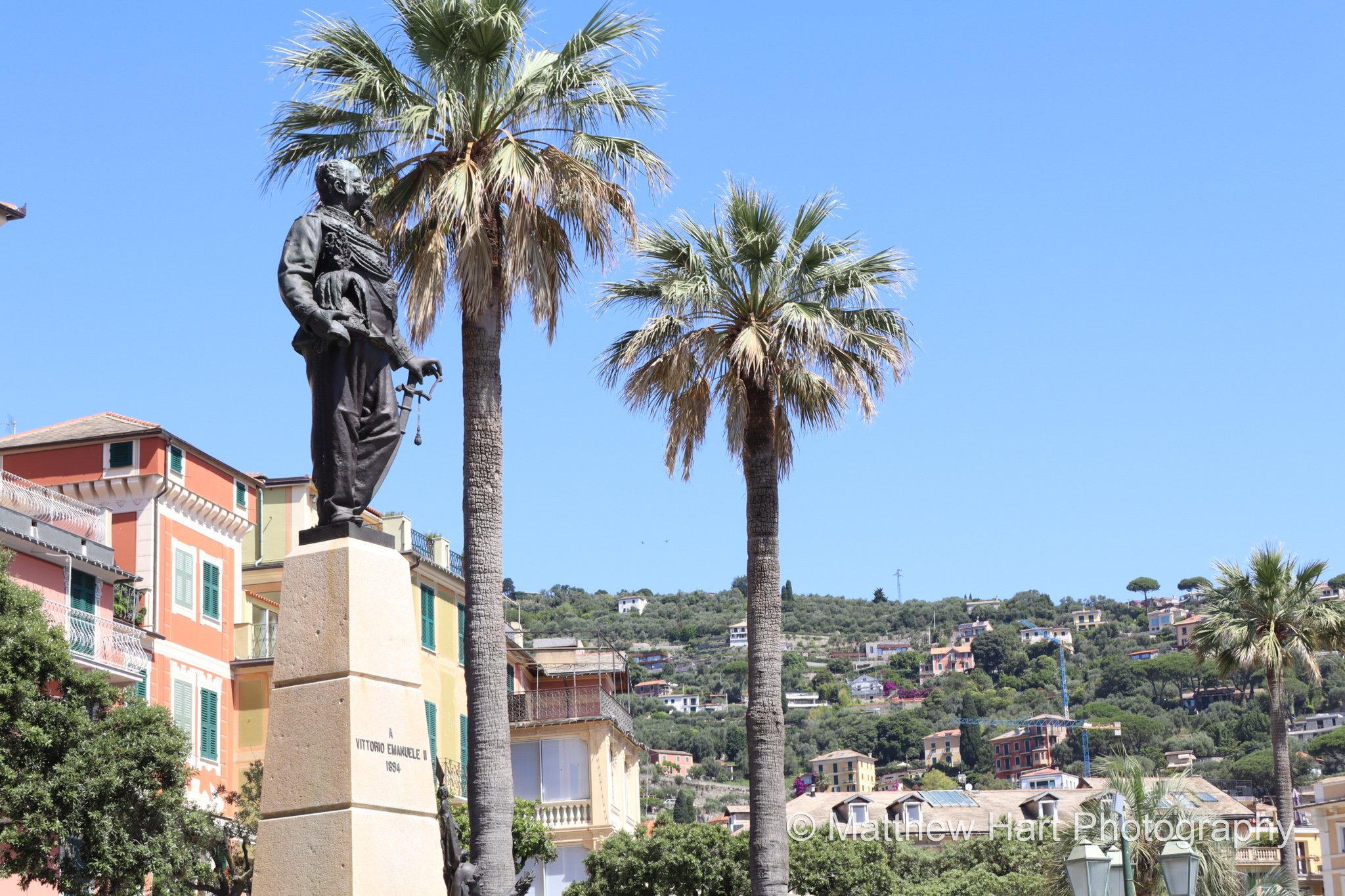 Fine art photograph of the bronze Vittorio Emanuele II statue between tall palm trees in Santa Margherita Ligure, Italy, with colorful buildings and hills behind.