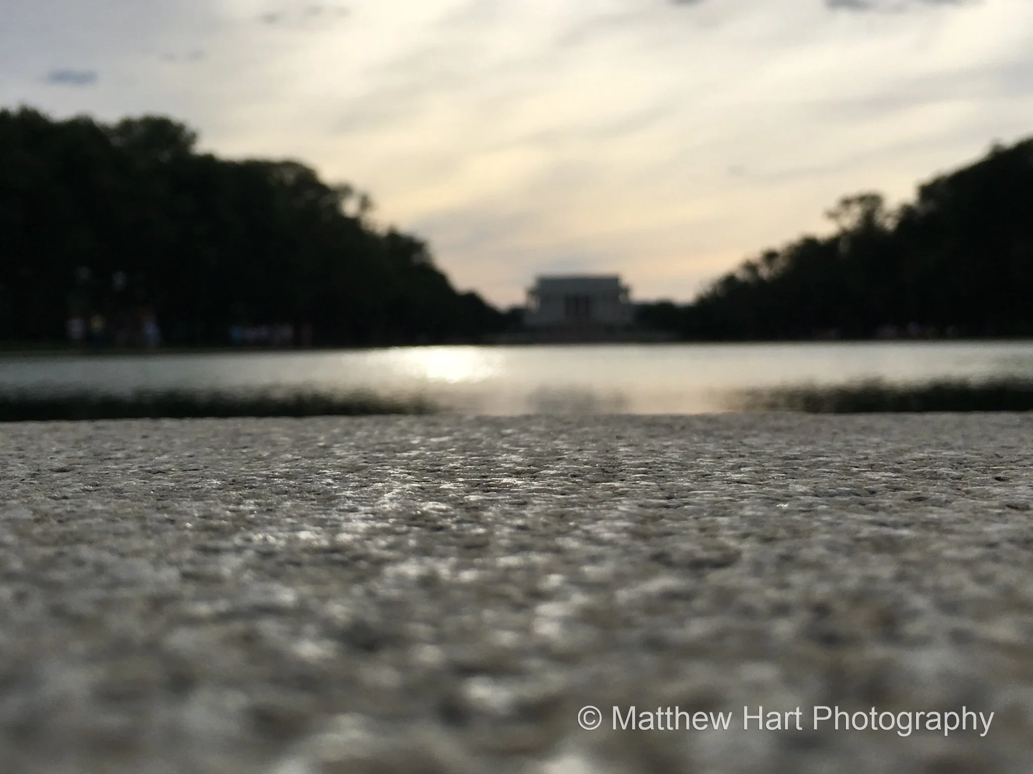 Fine art photograph from the granite edge of the Reflecting Pool looking toward the Lincoln Memorial between dark tree lines in Washington, D.C.