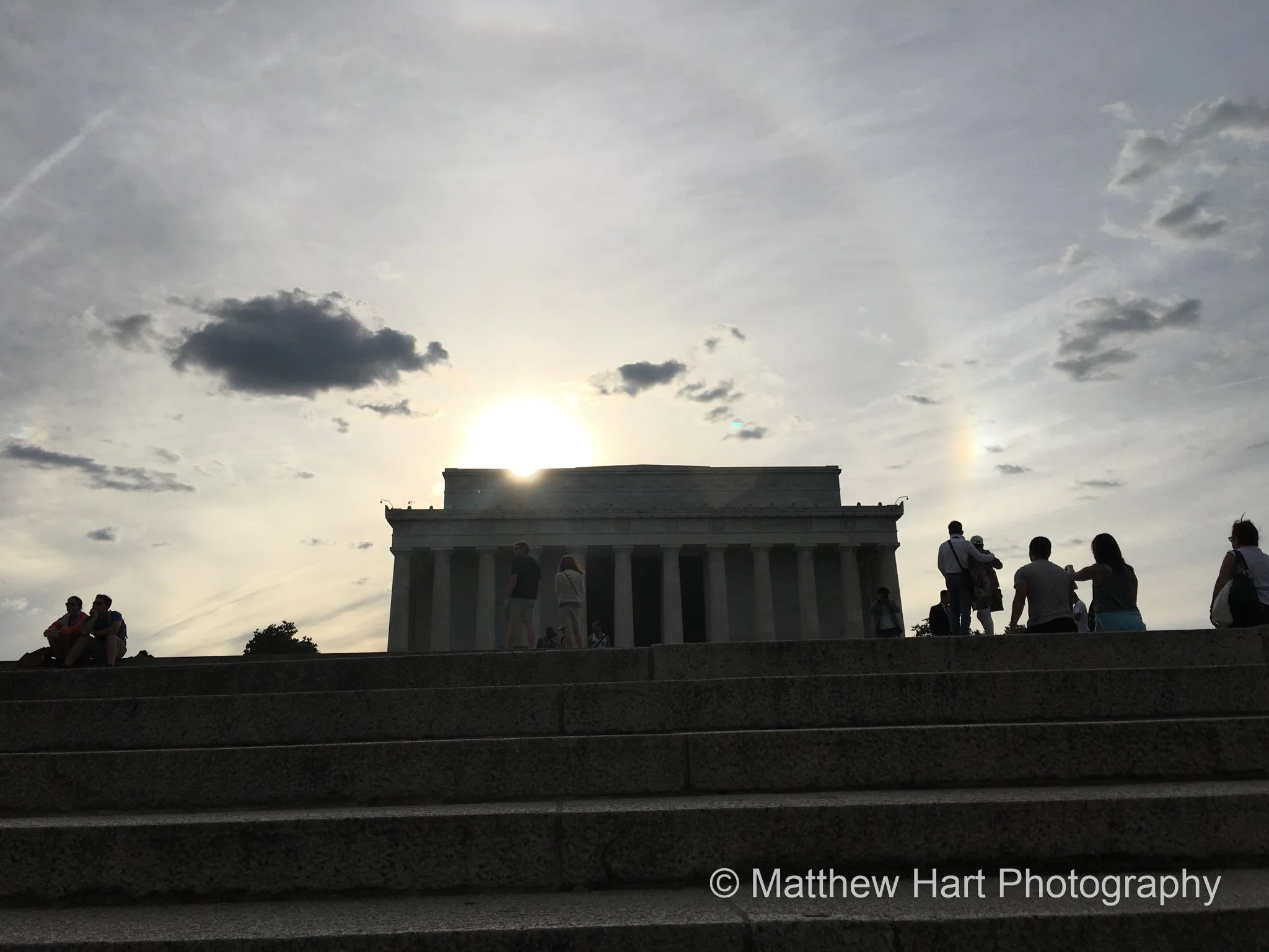 Fine art photograph of the Lincoln Memorial and small visitor silhouettes on the steps, backlit by the sun with a faint sundog in Washington, D.C.