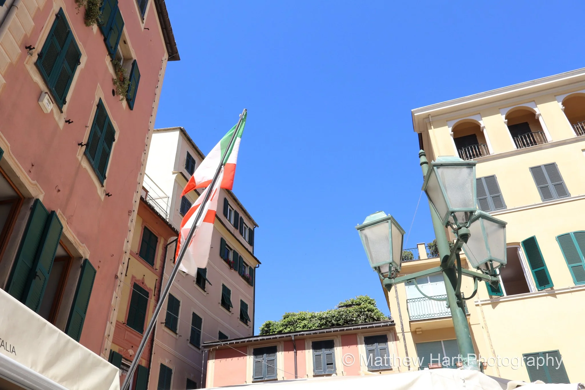 Fine art photograph looking up at an Italian flag between colorful buildings with green shutters and street lanterns in Santa Margherita Ligure, Italy.