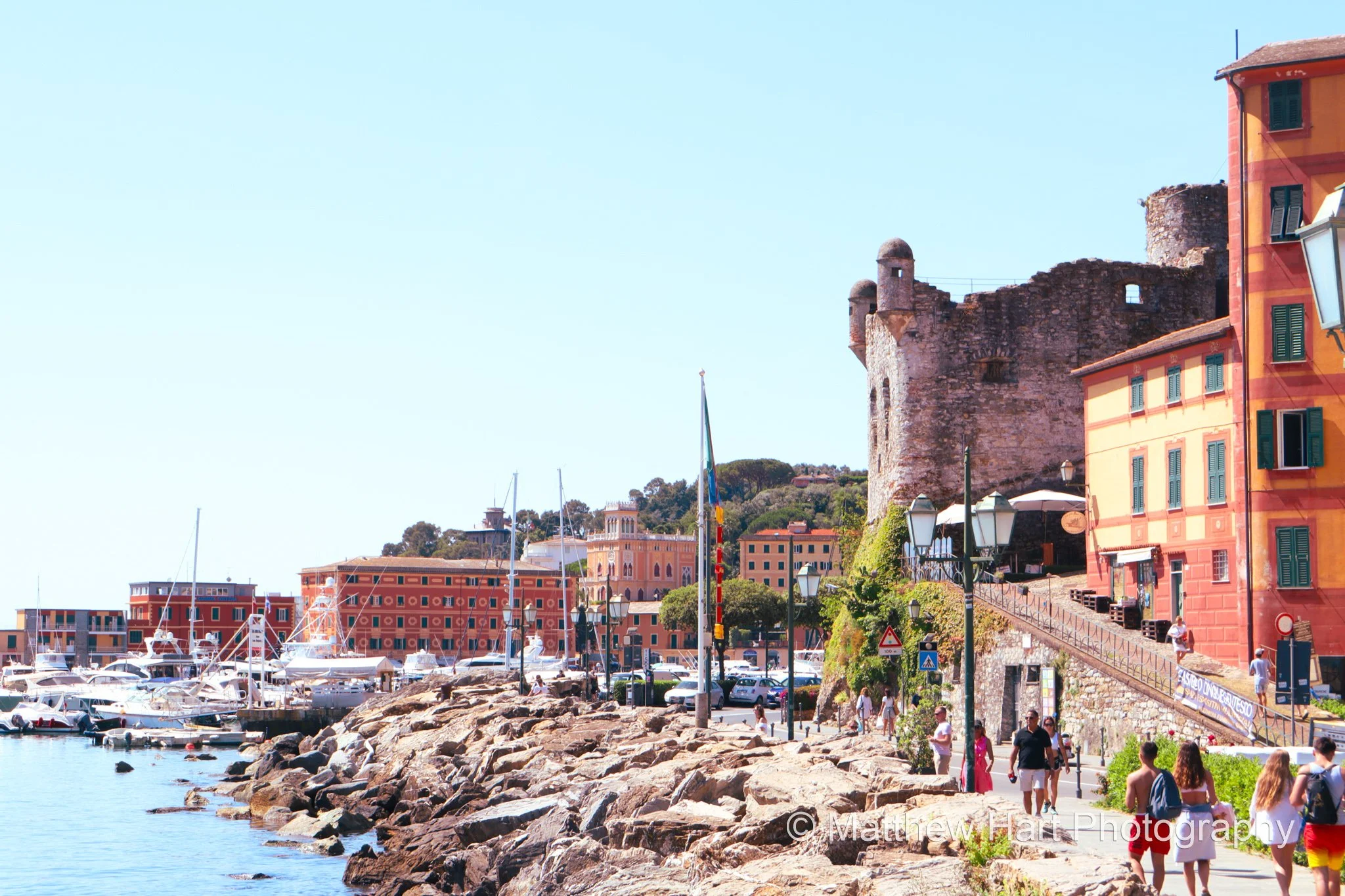 Fine art photograph of the stone castle at Santa Margherita Ligure, Italy, beside colorful townhouses, a rocky shoreline, and the harbor.