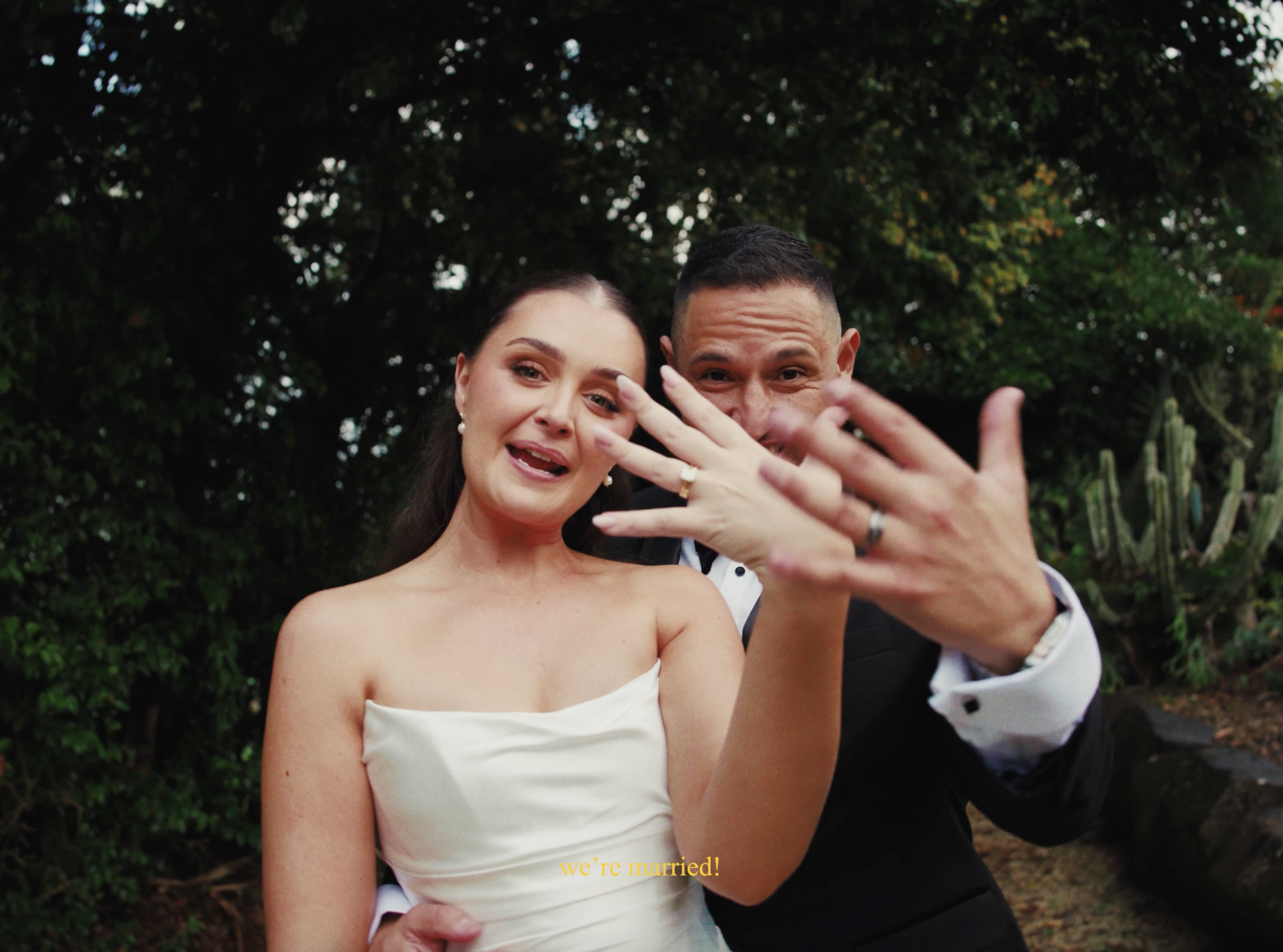 A woman in an elegant strapless dress and a man in a suit are celebrating outdoors, displaying their wedding rings, with the woman speaking and the man smiling behind her. The scene is lush with greenery in the background, and the text 'we're married!' is overlaid at the bottom.