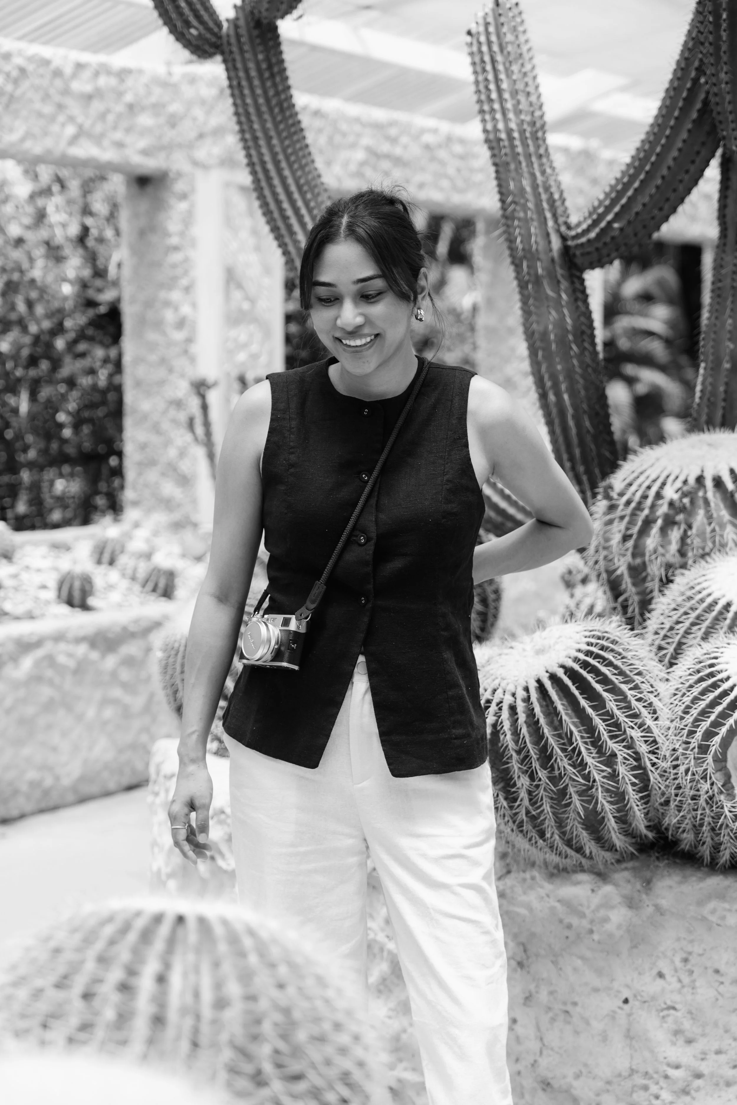 A woman smiling and looking down in front of large cacti, wearing a sleeveless black top and white pants, with a camera hanging around her neck.
