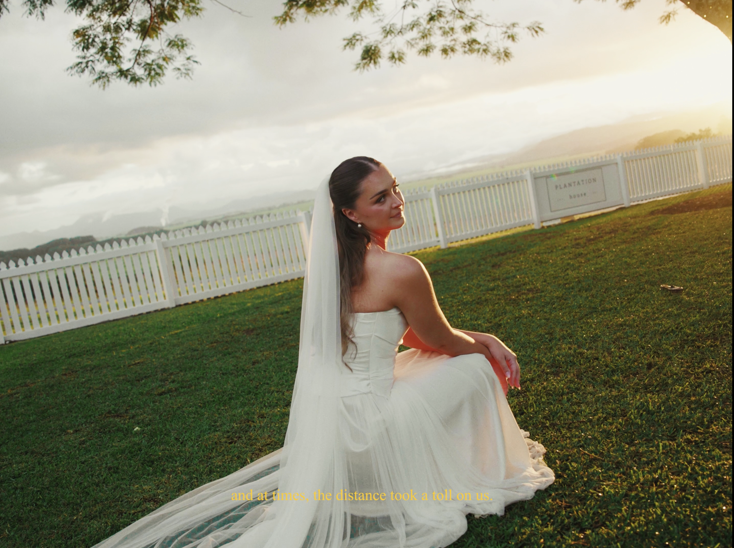 A woman in a white strapless wedding dress is sitting on the grass near a white picket fence, with trees and a cloudy sky in the background during sunset.