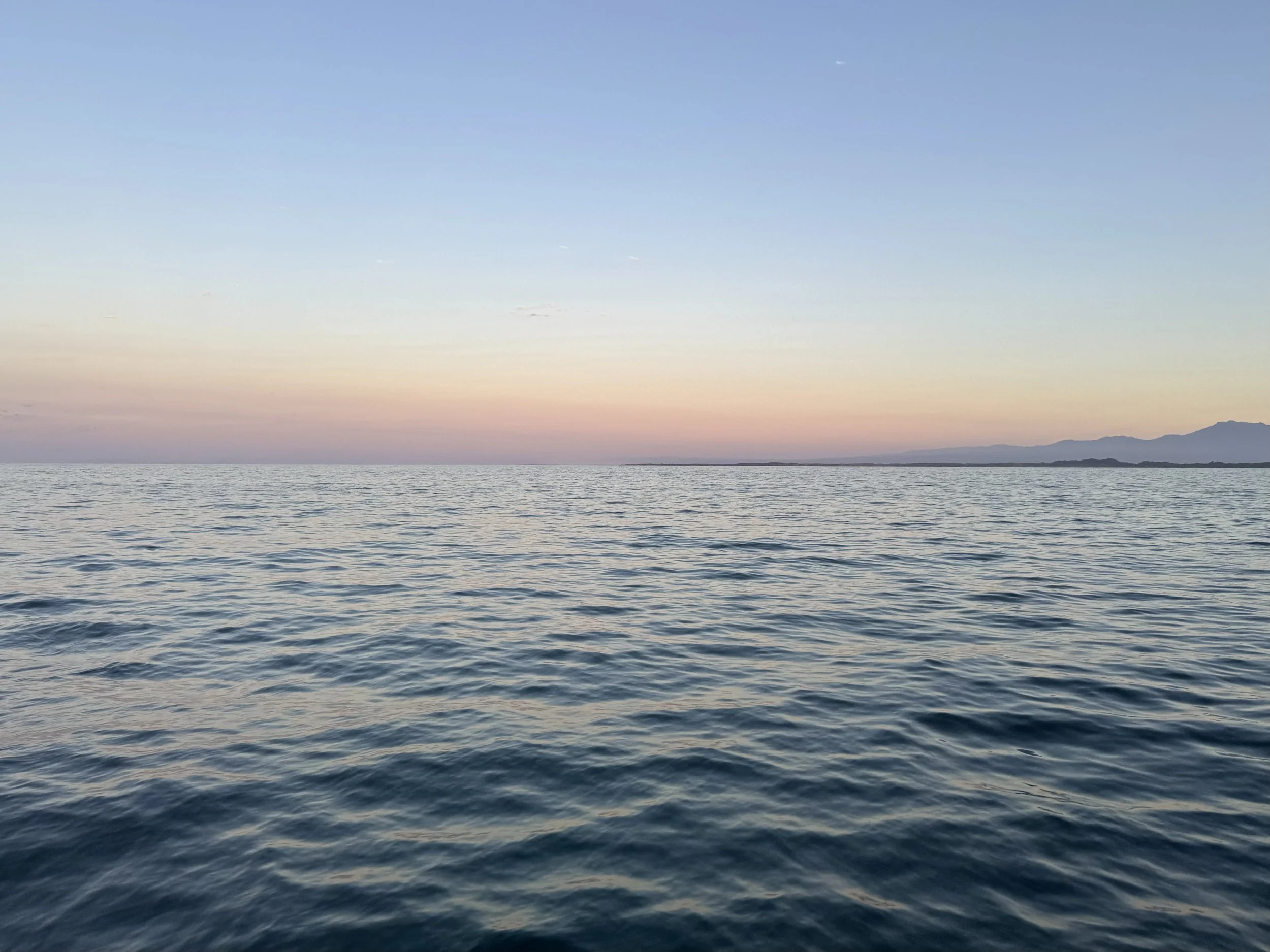 Calm ocean water with gentle waves during sunset, with mountain silhouette on the horizon and pastel-colored sky.