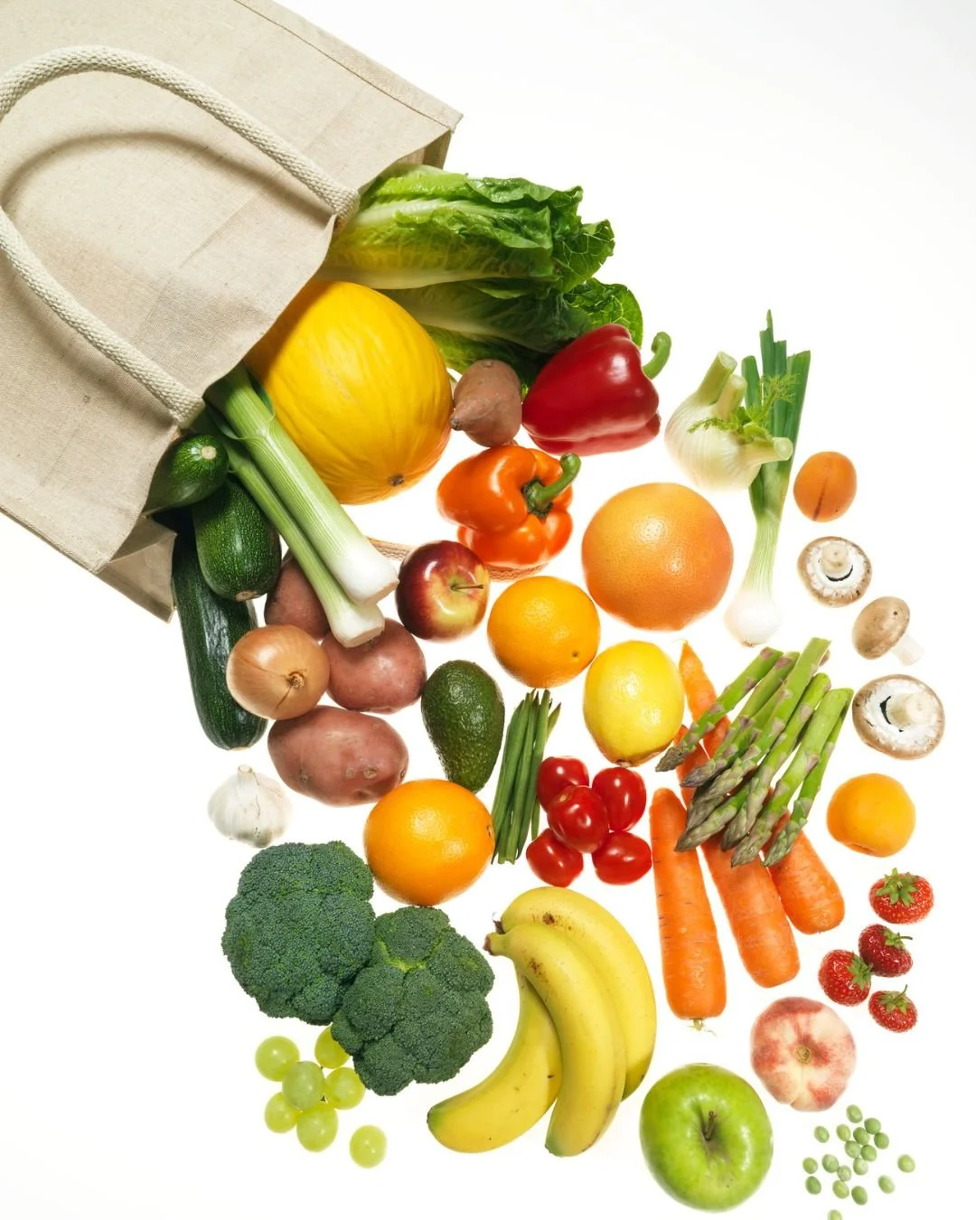 Assorted fresh fruits and vegetables spilling out of a beige reusable shopping bag, including broccoli, bananas, strawberries, grapes, carrots, asparagus, cherry tomatoes, apples, oranges, lemon, lime, avocado, onion, garlic, mushroom, and bell peppers.