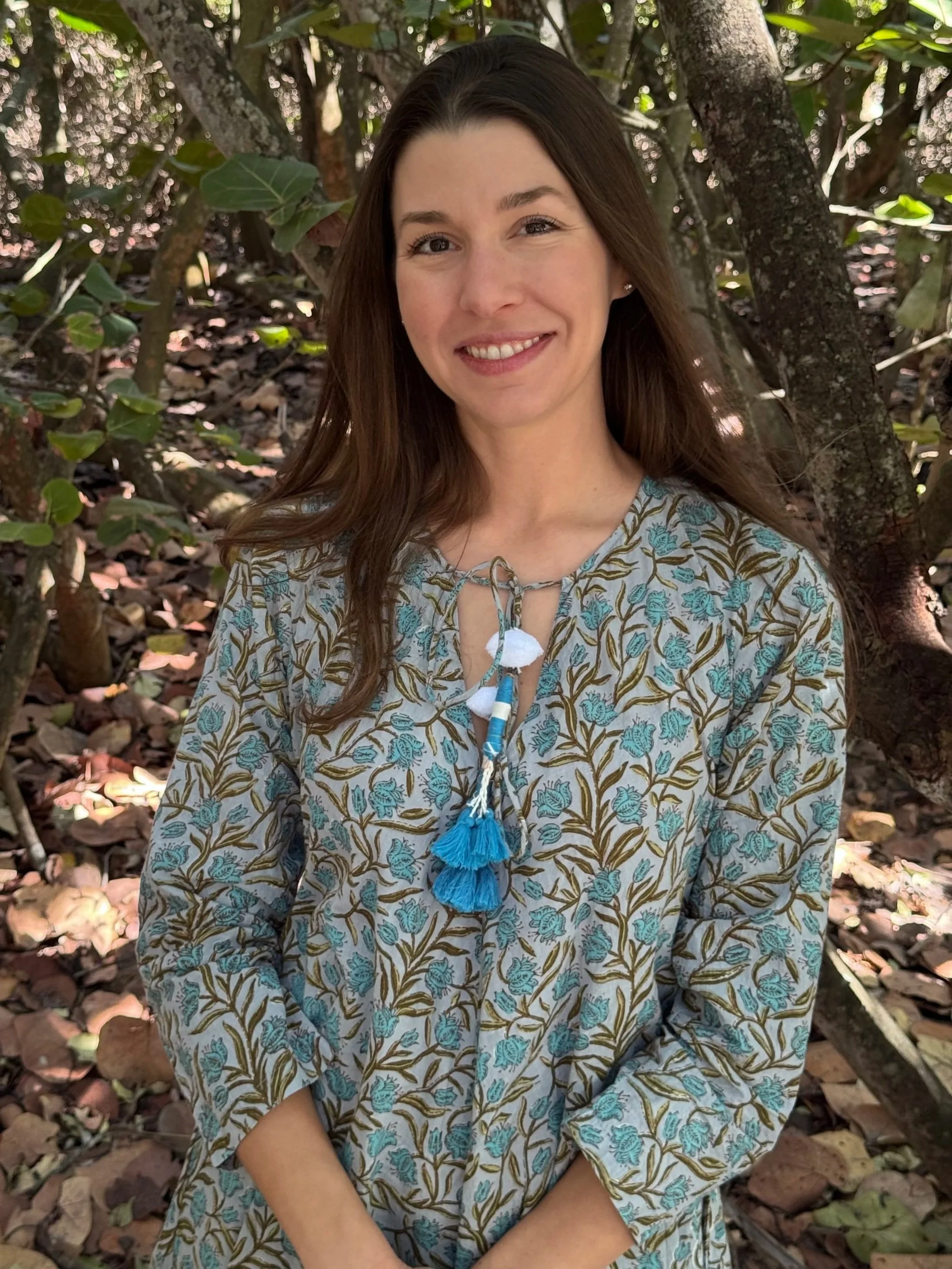 A woman with long brown hair smiling outdoors in a wooded area, wearing a patterned dress with blue and brown floral designs and a decorative necklace with blue tassels.