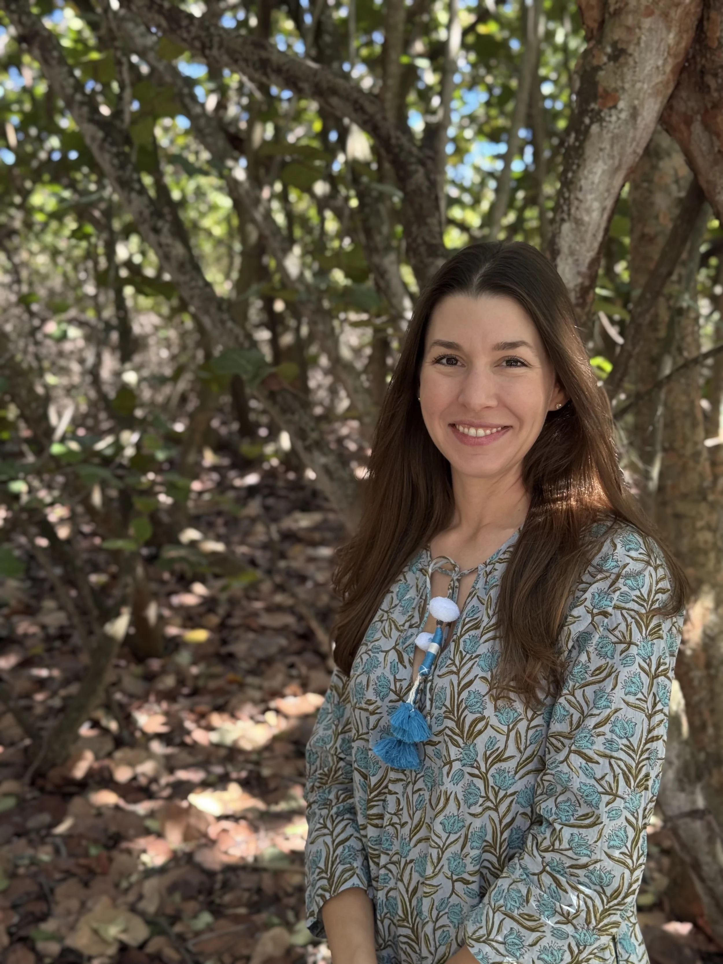 A woman standing outdoors in a leafy forest, smiling at the camera, wearing a patterned blouse with blue tassels on the front.