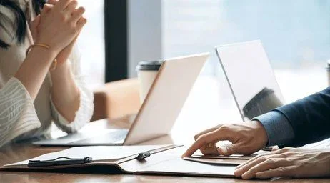 Two people sitting at a table using laptops, with a window in the background.