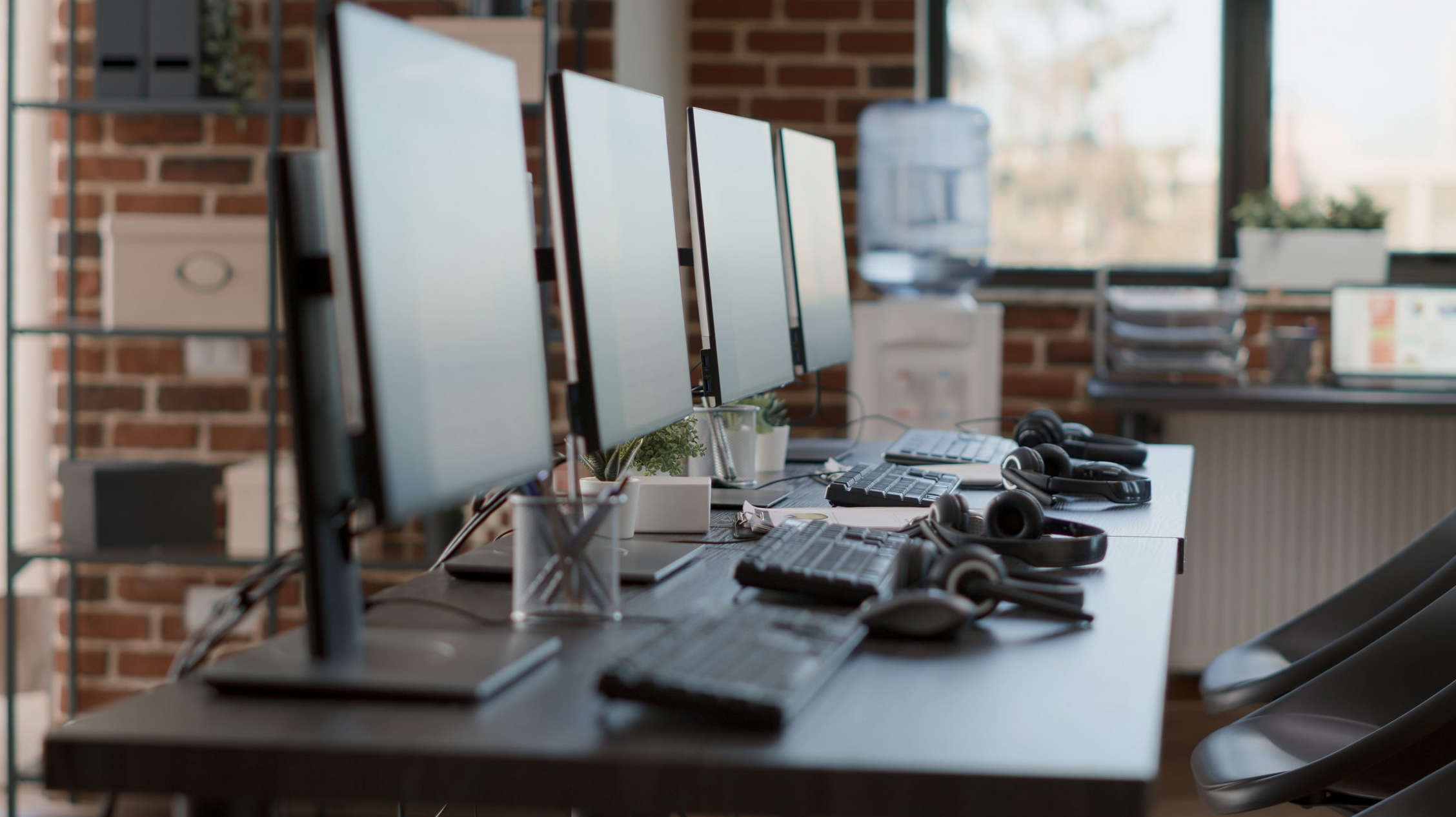 Office workspace with four computer monitors, keyboards, and headphones on a desk, with a brick wall and large window in the background.