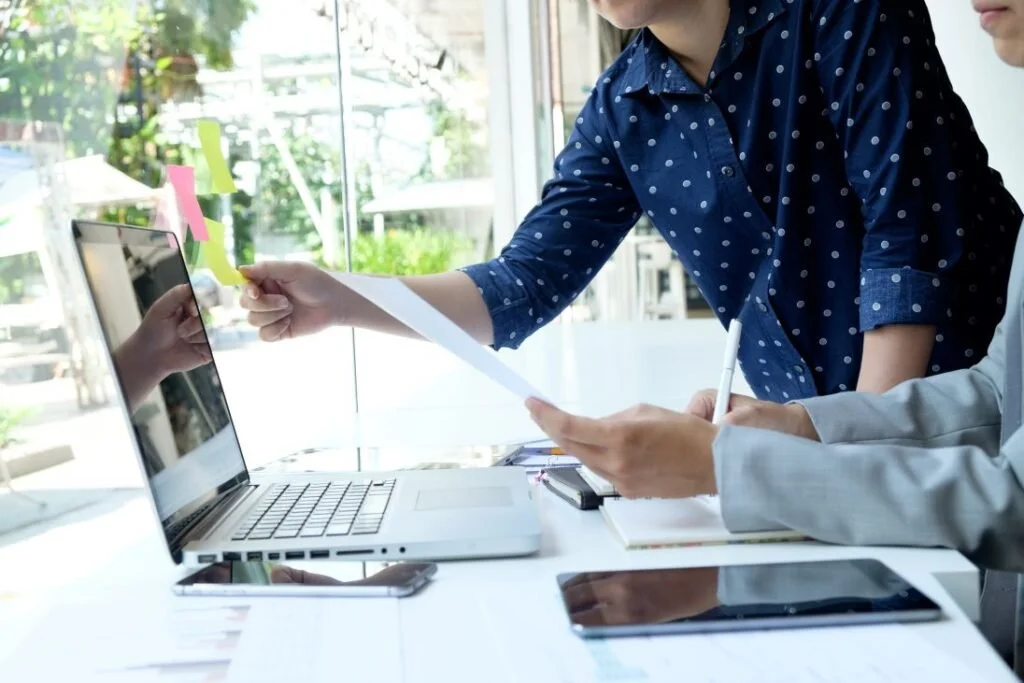 Two people collaborating at a desk with a laptop, smartphone, and tablet, exchanging documents in a bright office with large windows.
