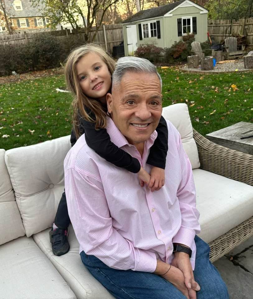 A young girl with long blonde hair smiling and giving a piggyback ride on an older man with gray hair, sitting on a beige outdoor sofa in a backyard with a grass lawn, trees, and a small shed in the background.